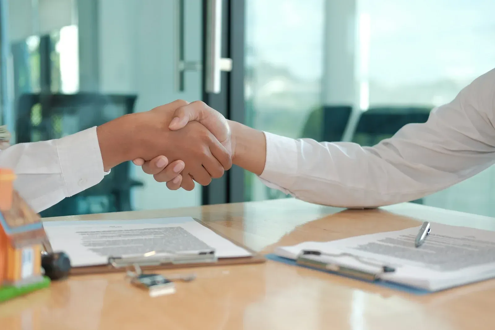 Two people shaking hands over a table with documents and a small house model; a deal closing.