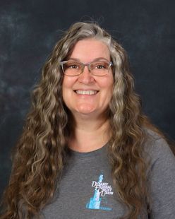 Woman with gray hair and glasses smiling, wearing a gray shirt with a logo, against a teal background.