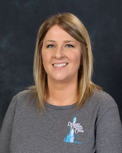Blonde woman smiling, wearing gray shirt with logo, against blue background.
