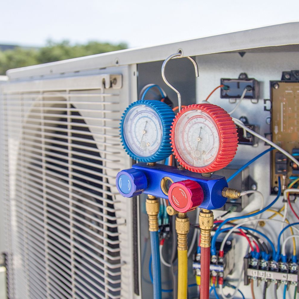 HVAC manifold gauges with blue and red dials attached to the open service panel of an outdoor air conditioning unit.