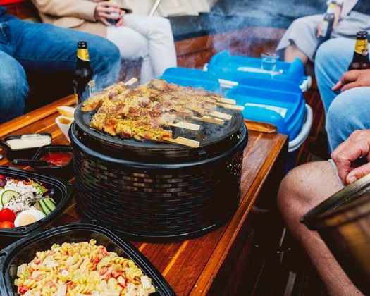 Mensen verzamelden zich rond een tafel met gegrild vlees, drankjes en bijgerechten voor een maaltijd in de buitenlucht op een boot.