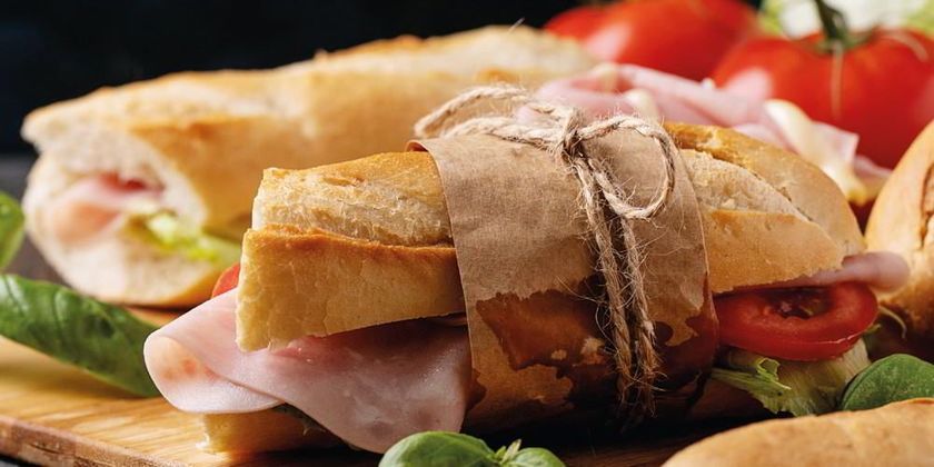 Een ingepakte sandwich met ham, sla en tomaat op een rustieke houten plank.