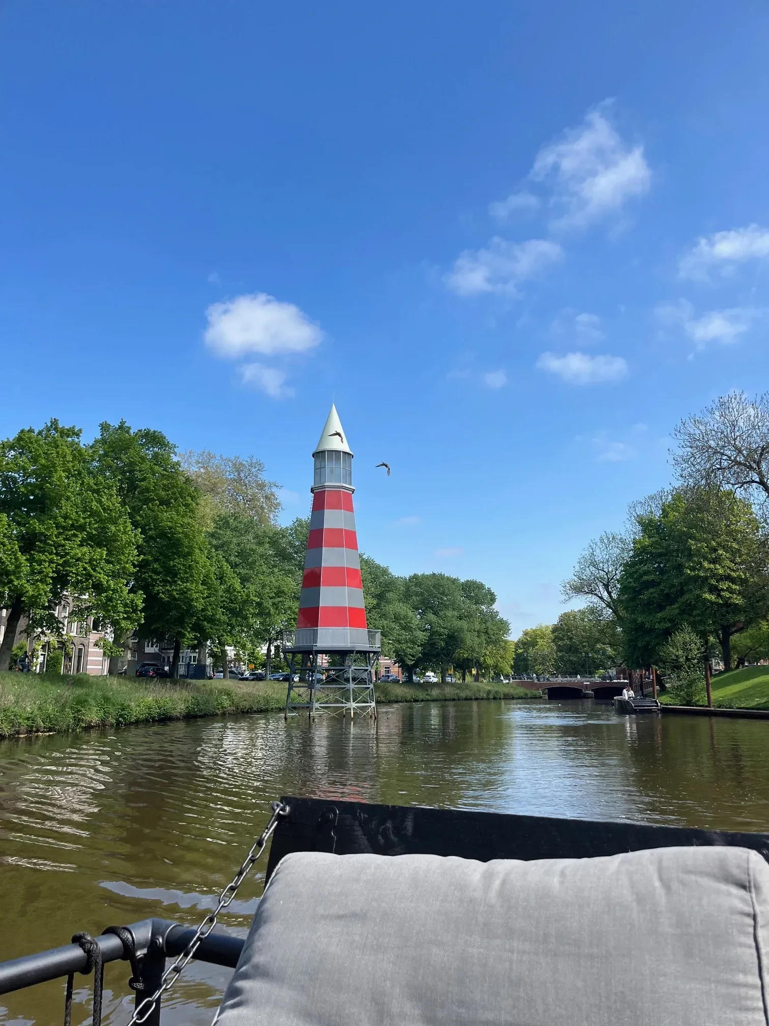 Een rood-wit gestreepte vuurtoren in een Breda. Groene bomen staan ​​langs de waterkant onder een blauwe hemel.