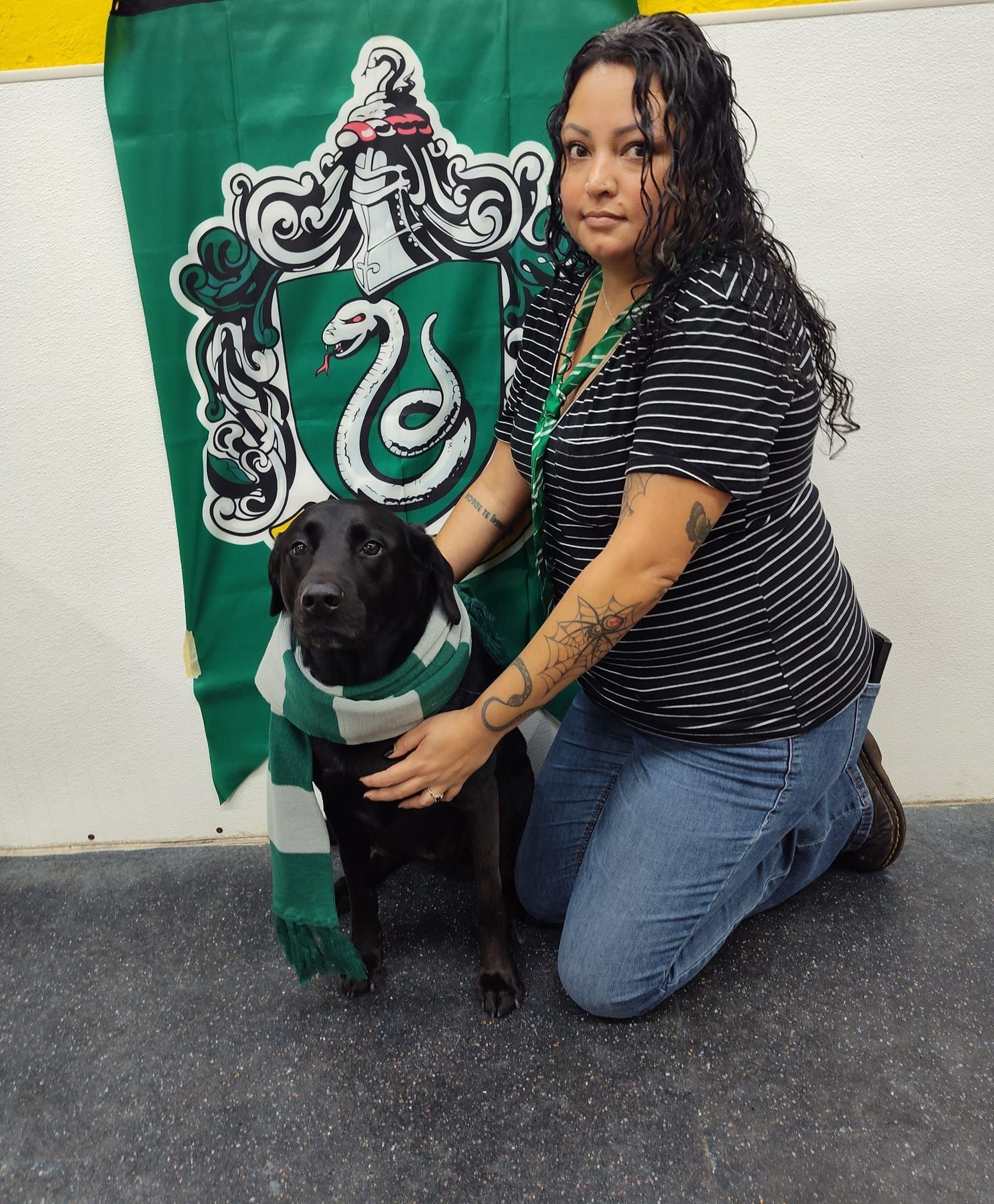 A woman is kneeling next to a black dog wearing a slytherin scarf