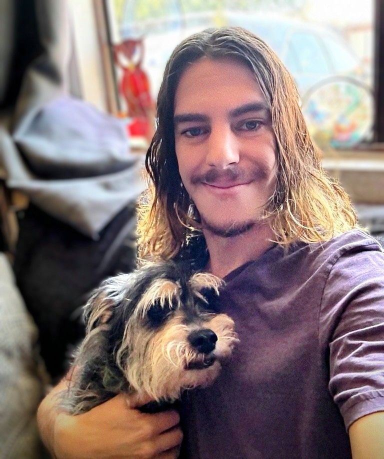 A man with long hair is holding a small dog in his arms.