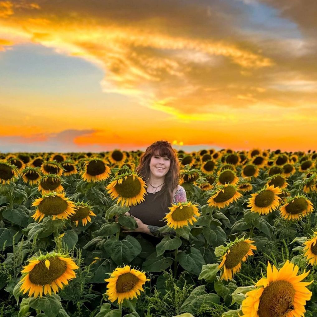 A woman is standing in a field of sunflowers at sunset.