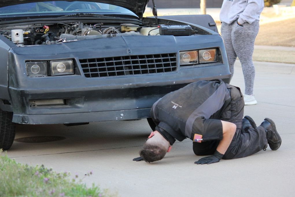 A man is kneeling down under the hood of a car.