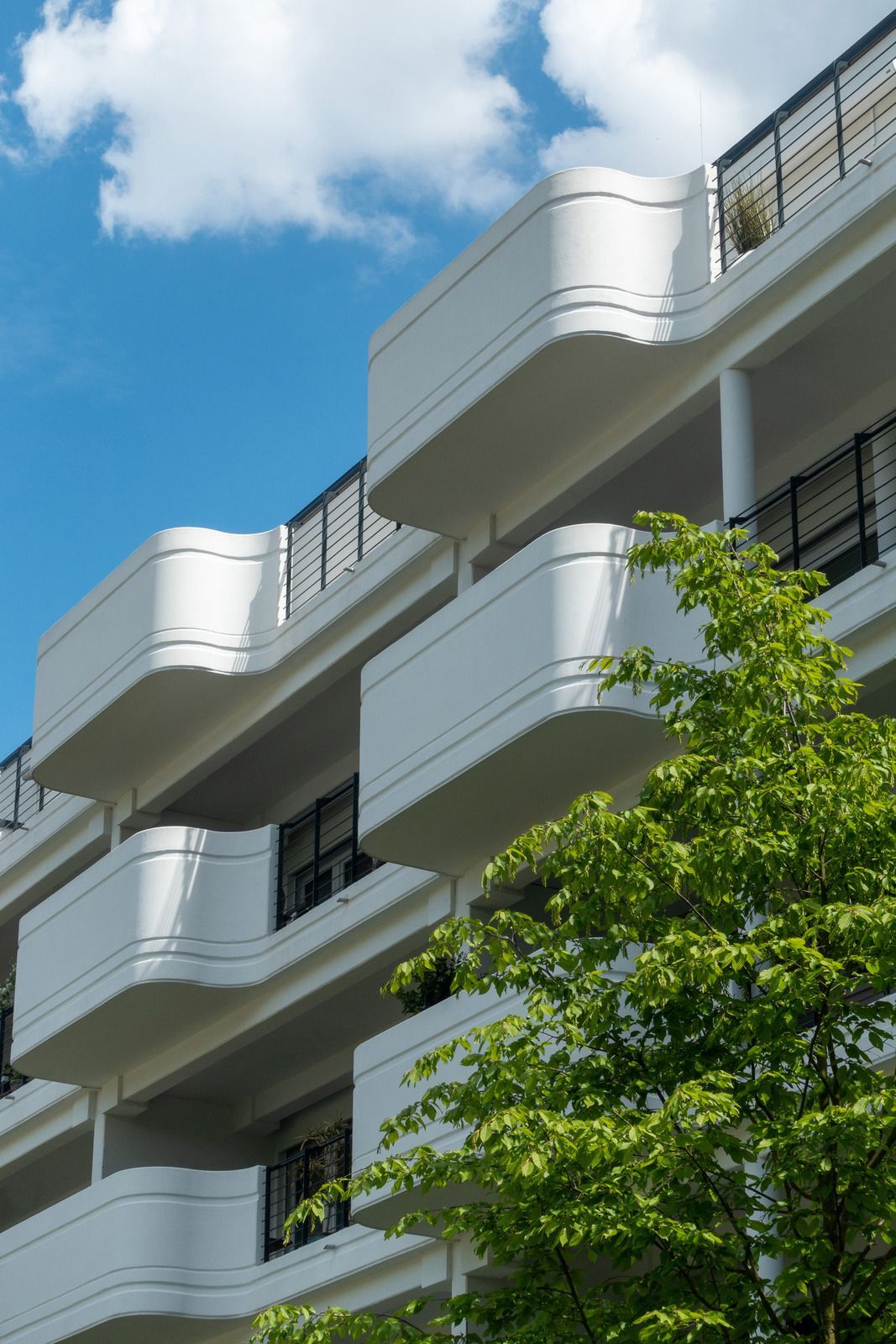 Modern white apartment building with curved balconies against a blue sky with clouds and a foreground tree.