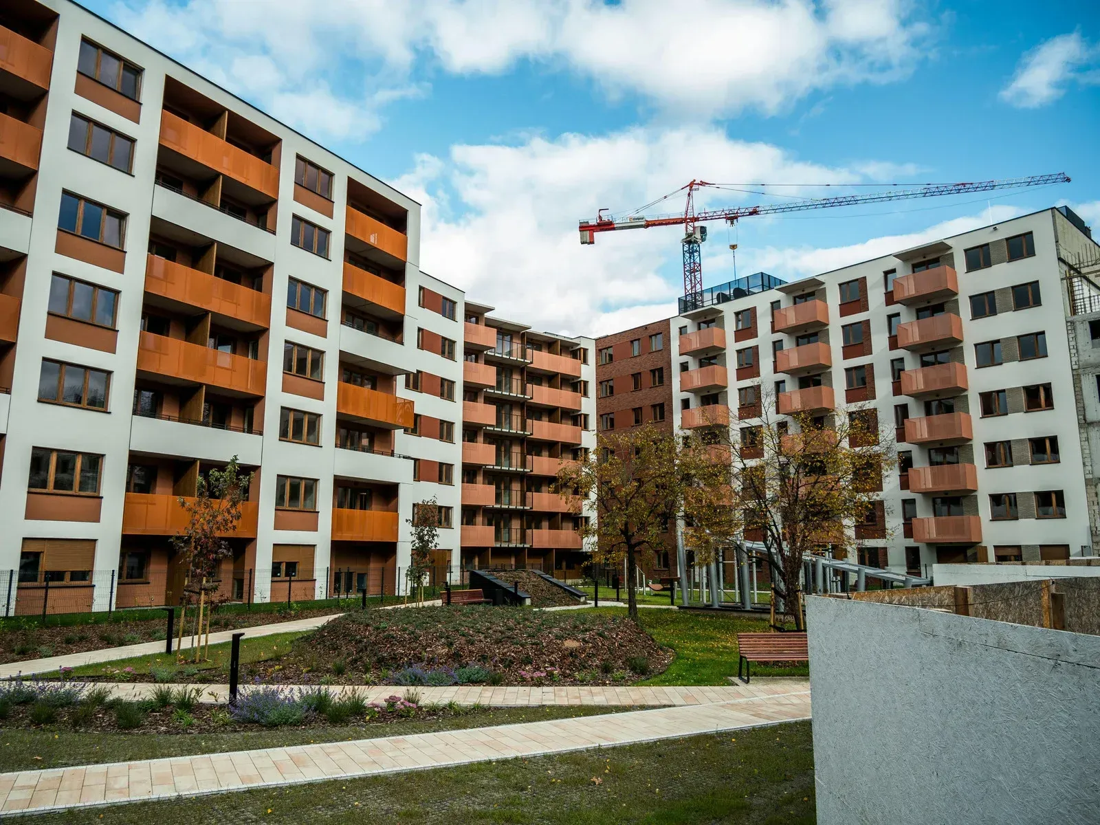 Modern apartment buildings with orange and white facades, green landscaping, and a construction crane against a blue sky.