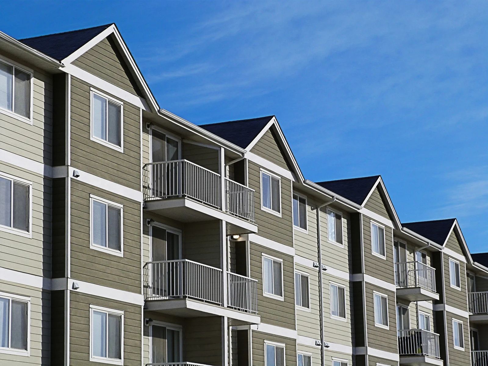 A multi-story apartment building with sage green siding, white trim, and balconies against a clear blue sky.