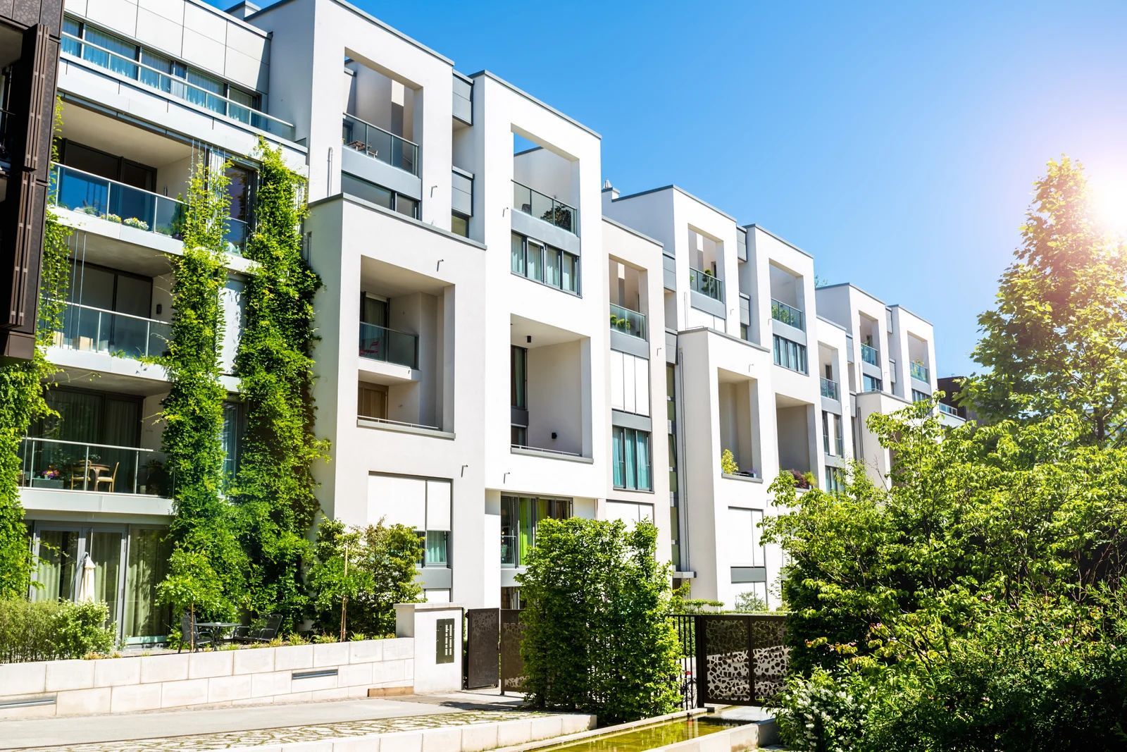 Modern white apartment building with balconies, lush greenery, and blue sky.