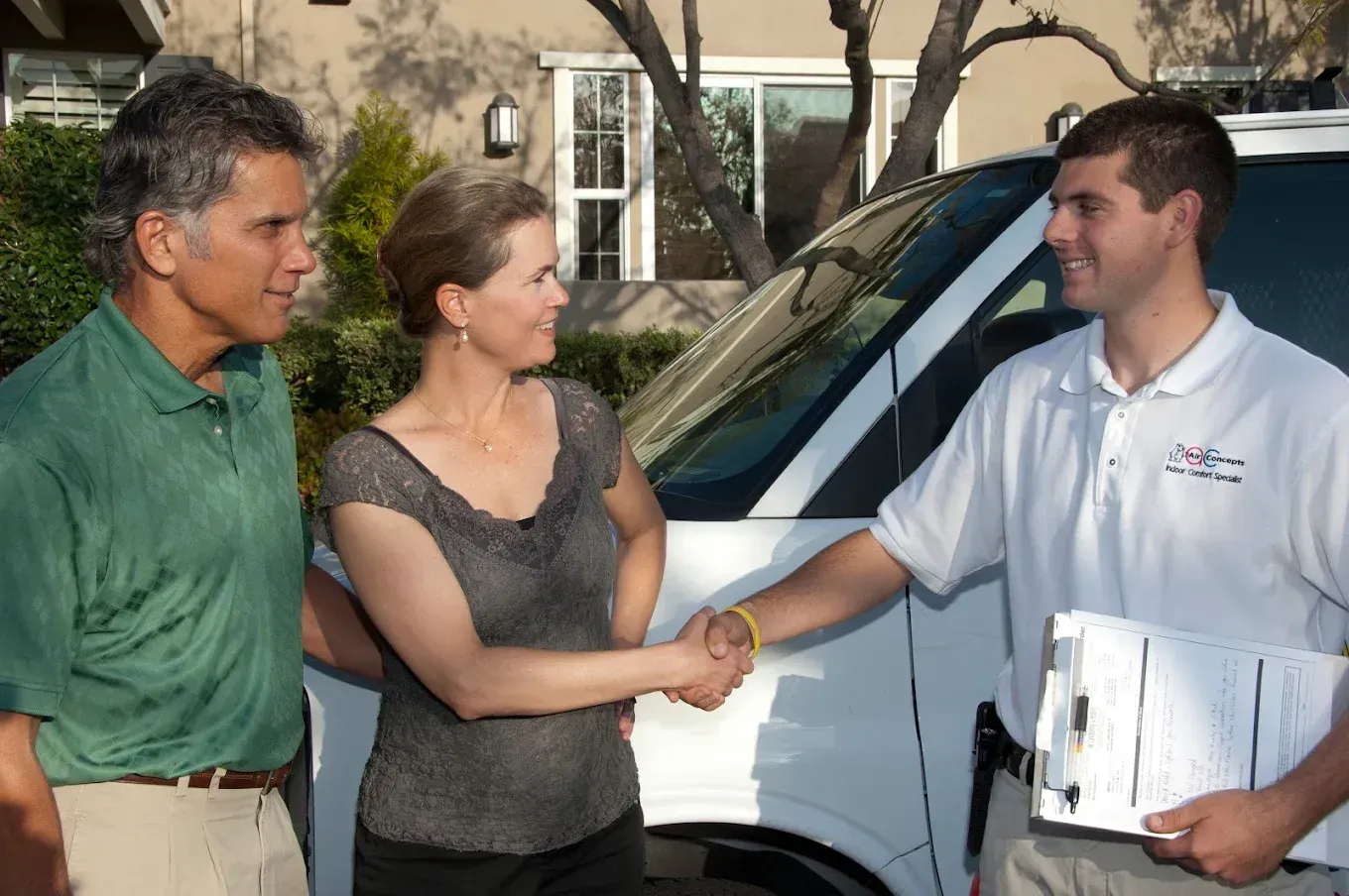 Man shaking hands with a woman, standing with a man by a white van in front of a house.
