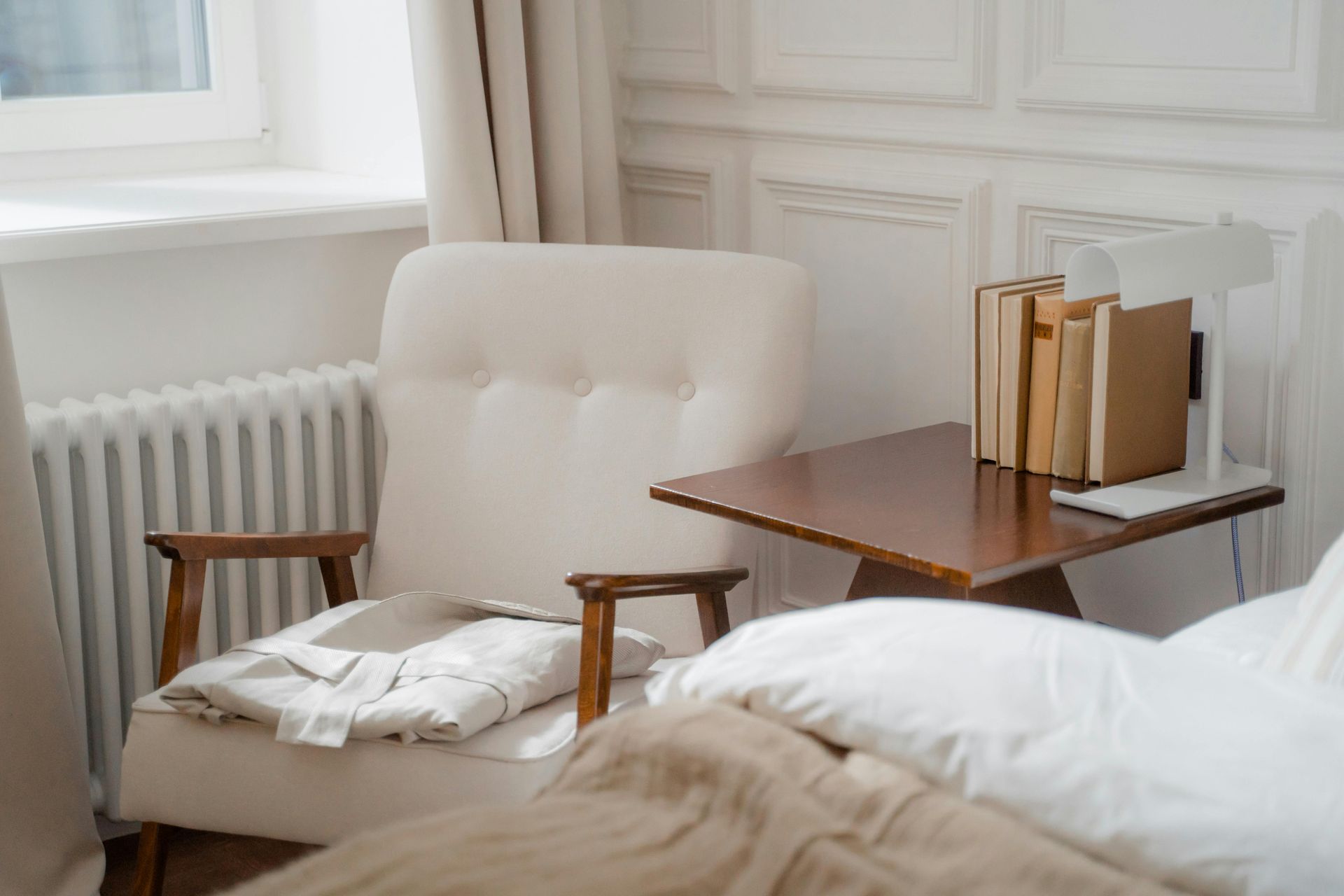 Cozy bedroom corner: beige armchair, brown side table with books, white lamp, and partially visible bed.