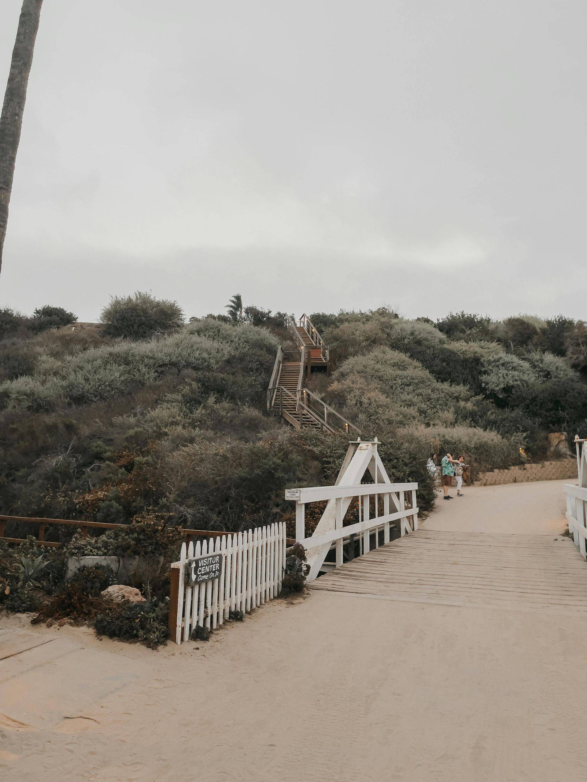 White gate and bridge leading to wooden stairs ascending a grassy hill under a cloudy sky.