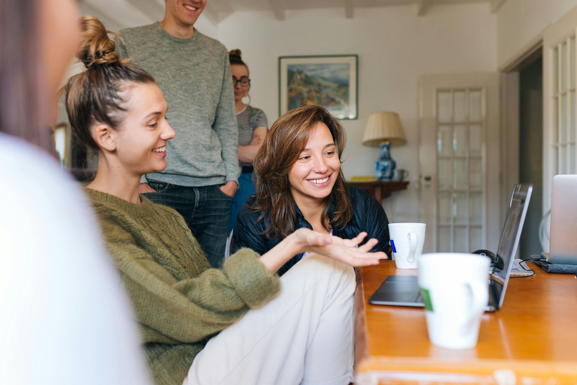 Group of people looking at a laptop, smiling.  One woman gestures with her hand; others watch. Indoors, with coffee cups.