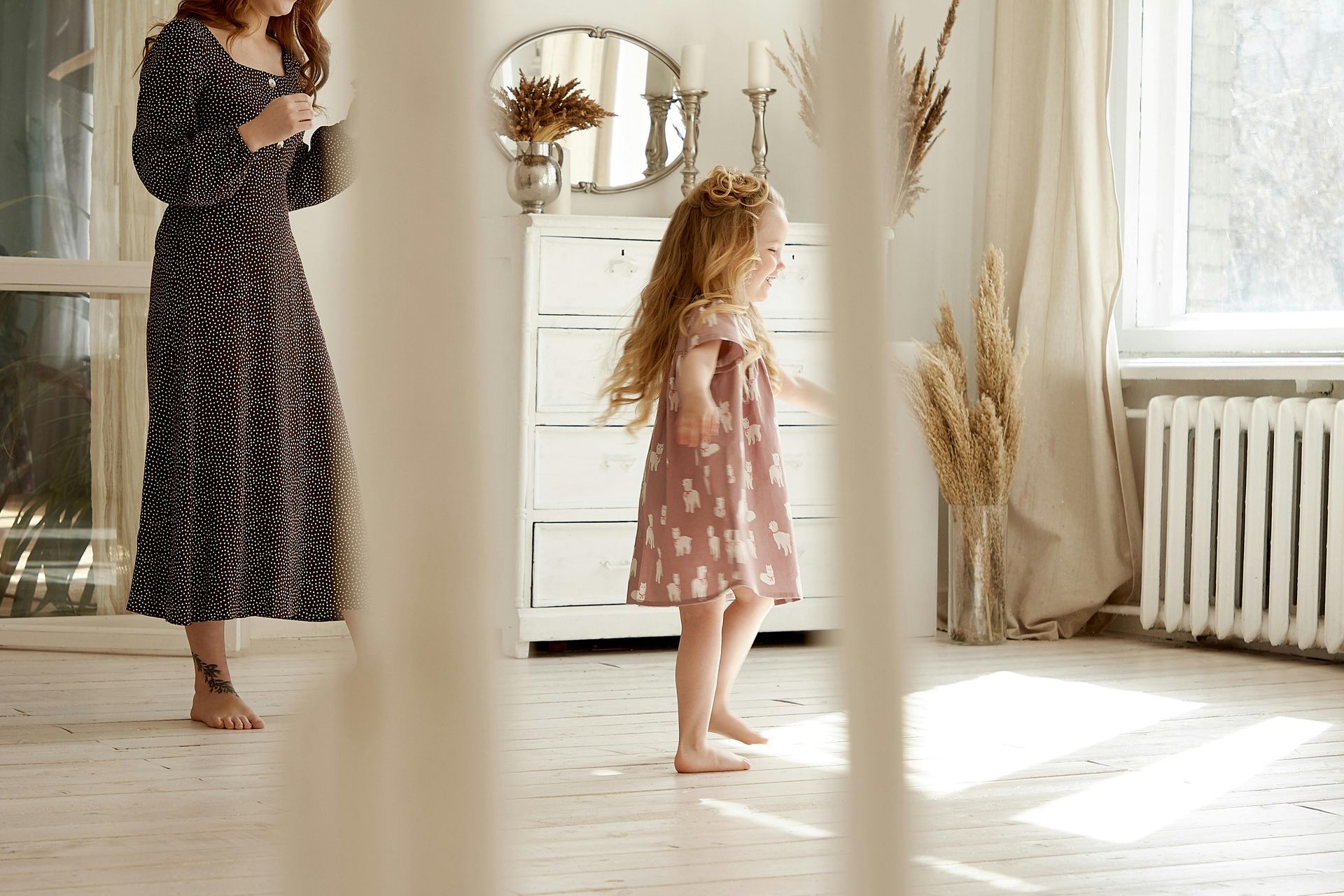 Woman in a black patterned dress looks at phone as barefoot girl in pink dress twirls in a sunny room.