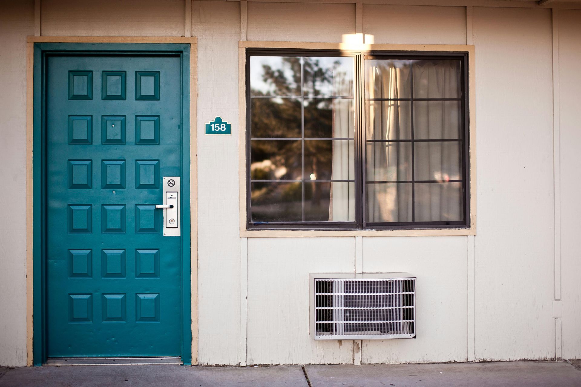 Teal door with window and air conditioner on the side of a cream-colored building.