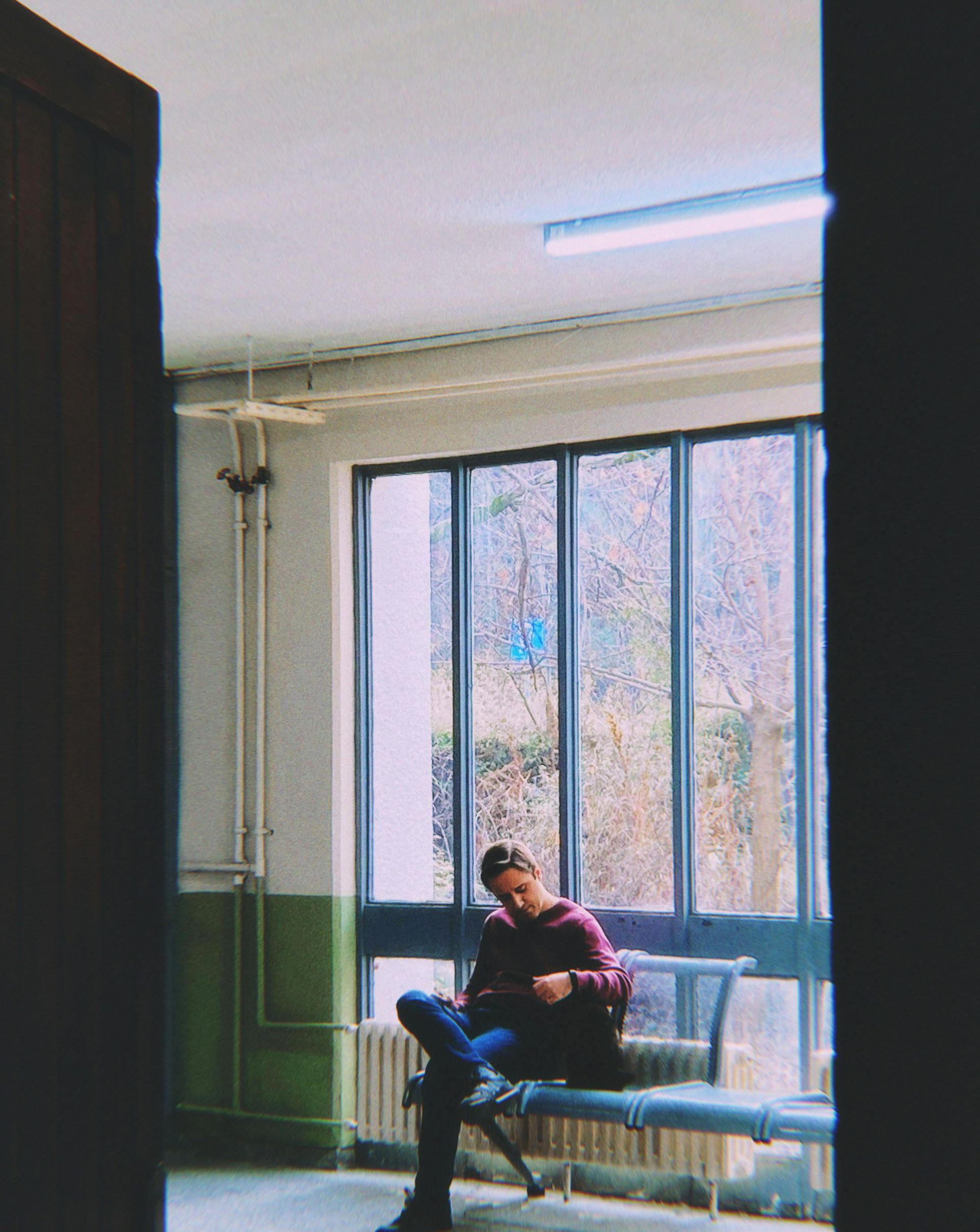 Person sits on a bench, looking at a device, in front of a window. Building interior with a view of trees.
