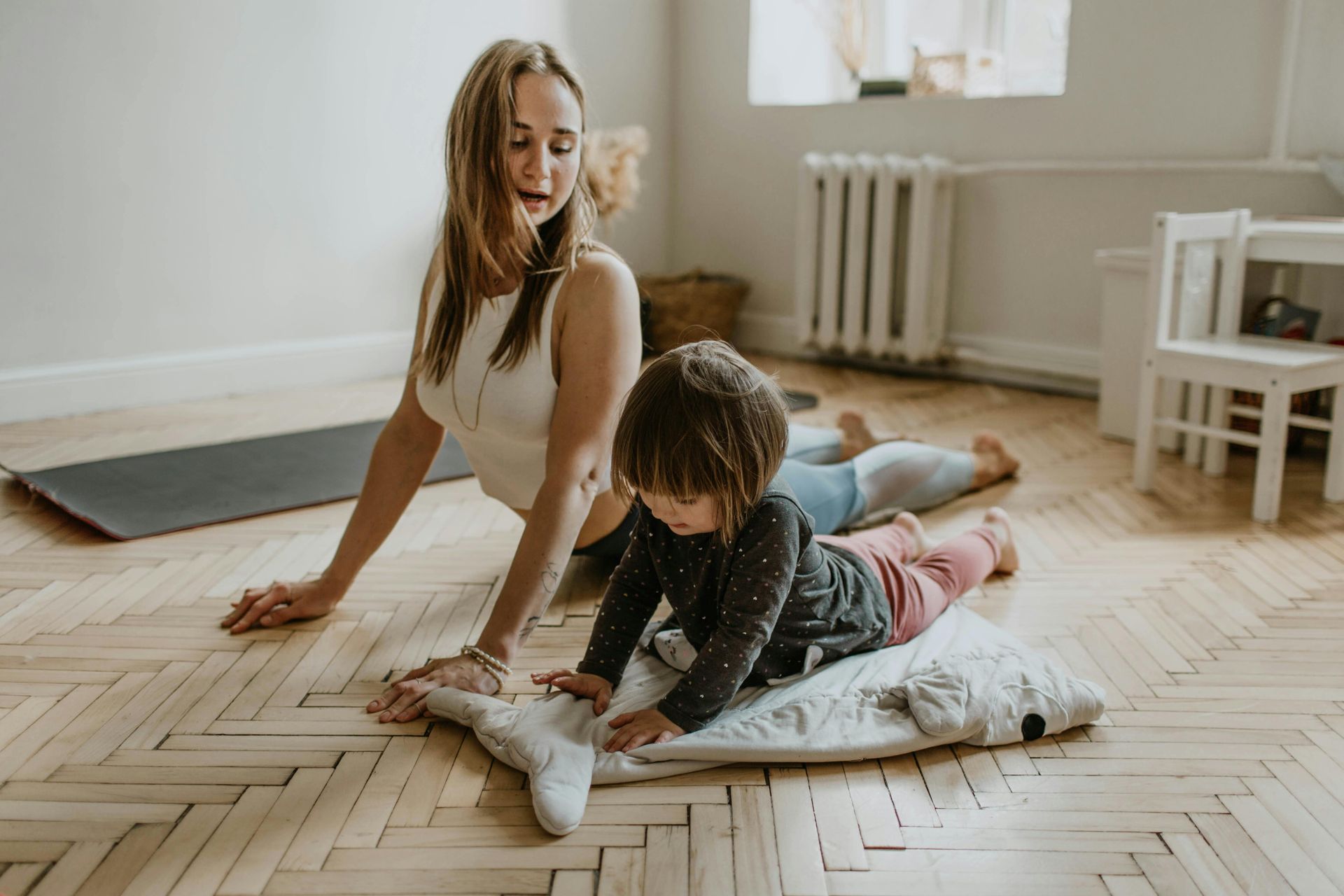 Woman and child practicing yoga on a wooden floor, indoor. A second child is visible in the background.