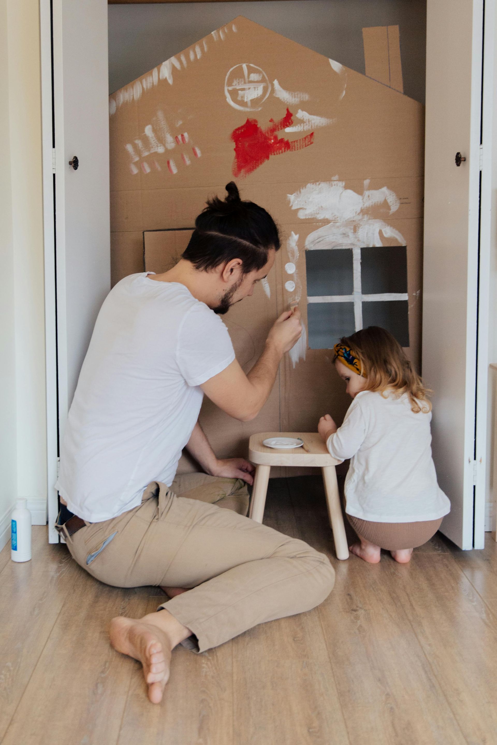Man and child paint cardboard house in a closet; white paint, tan pants, wooden stool.