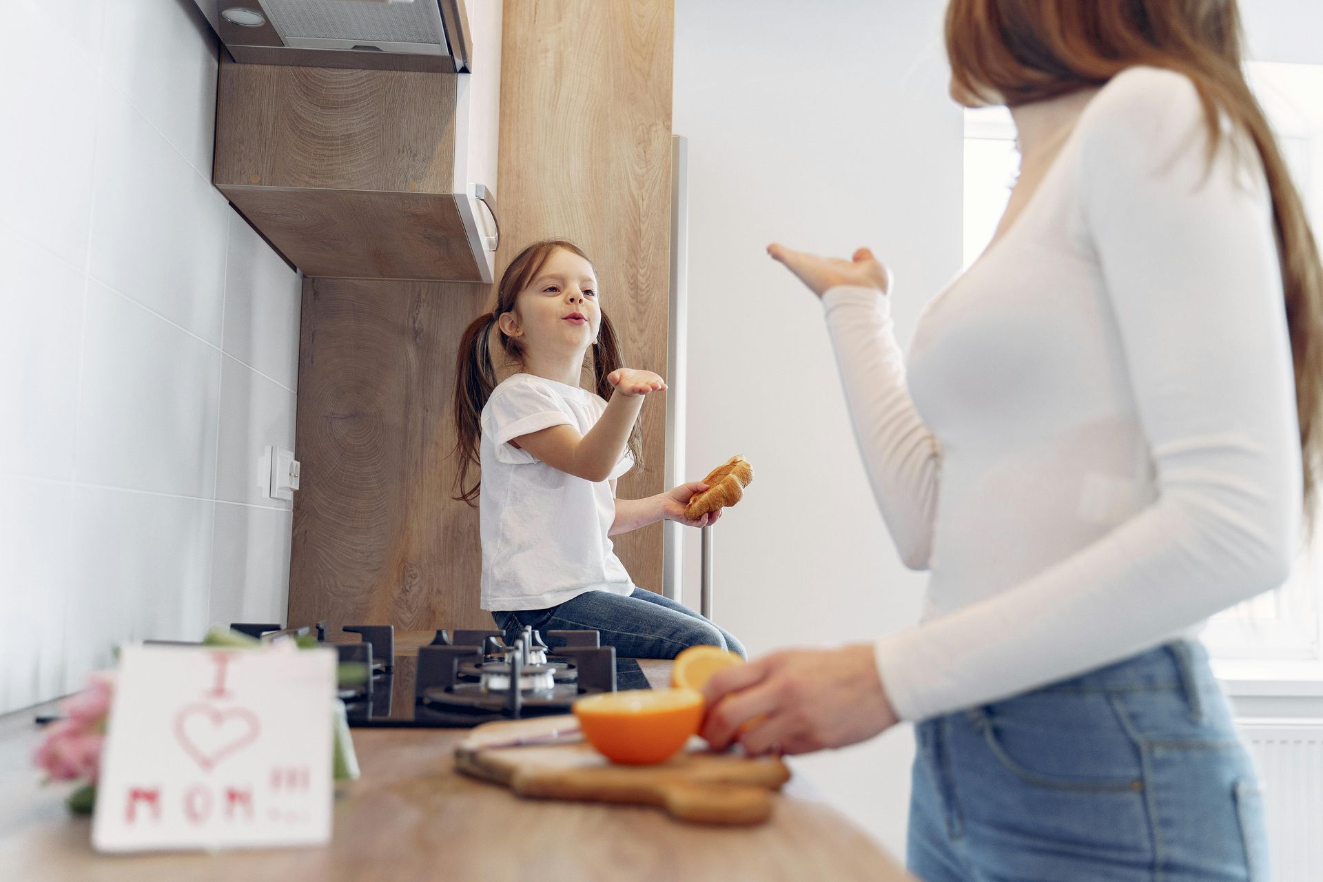 Mother and child in a kitchen, preparing food. Child sits on counter, holding orange. Mom holds orange and gestures.