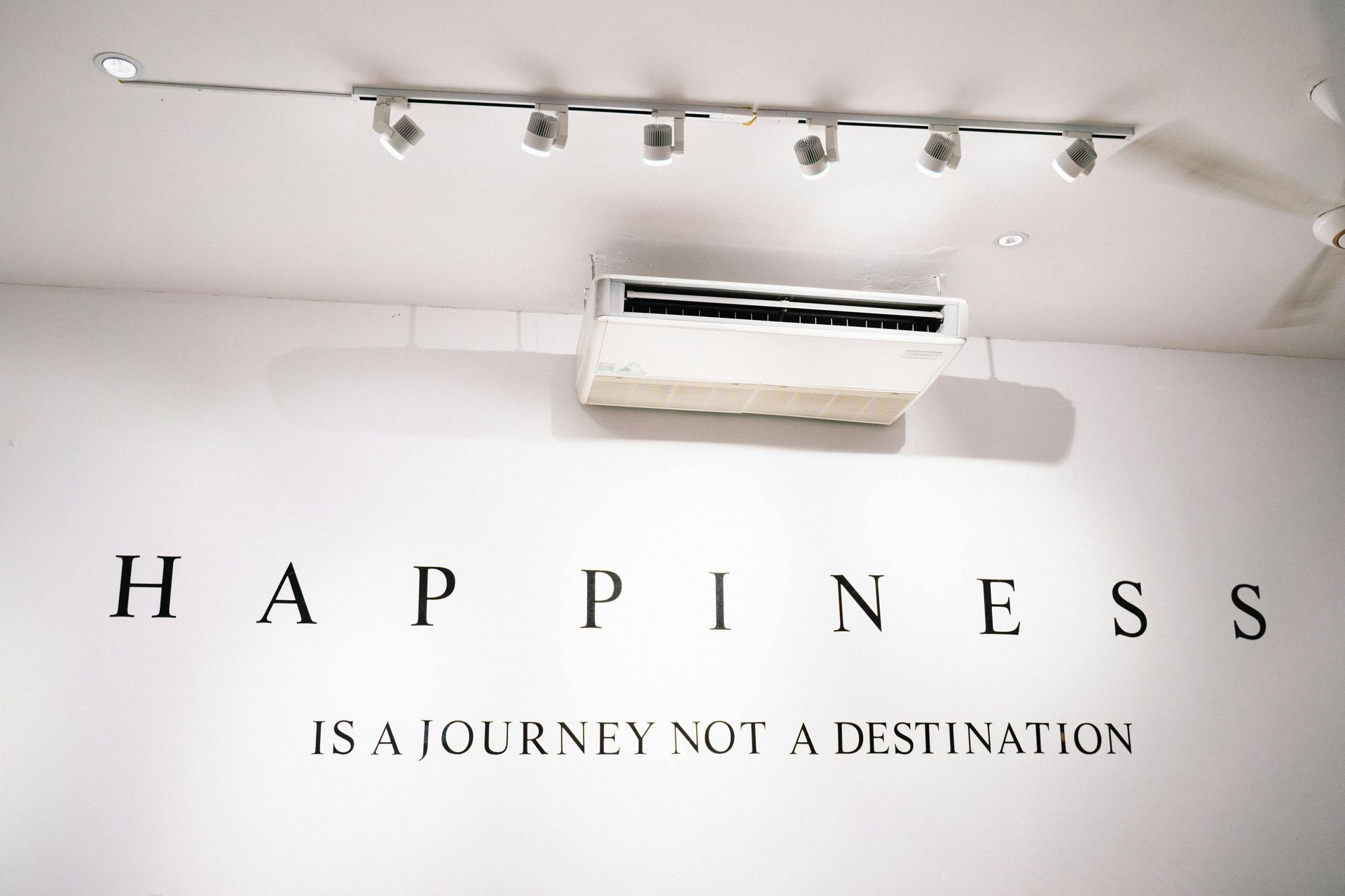 White wall with “Happiness is a journey not a destination” text. Above, a row of spotlights and an air conditioner.
