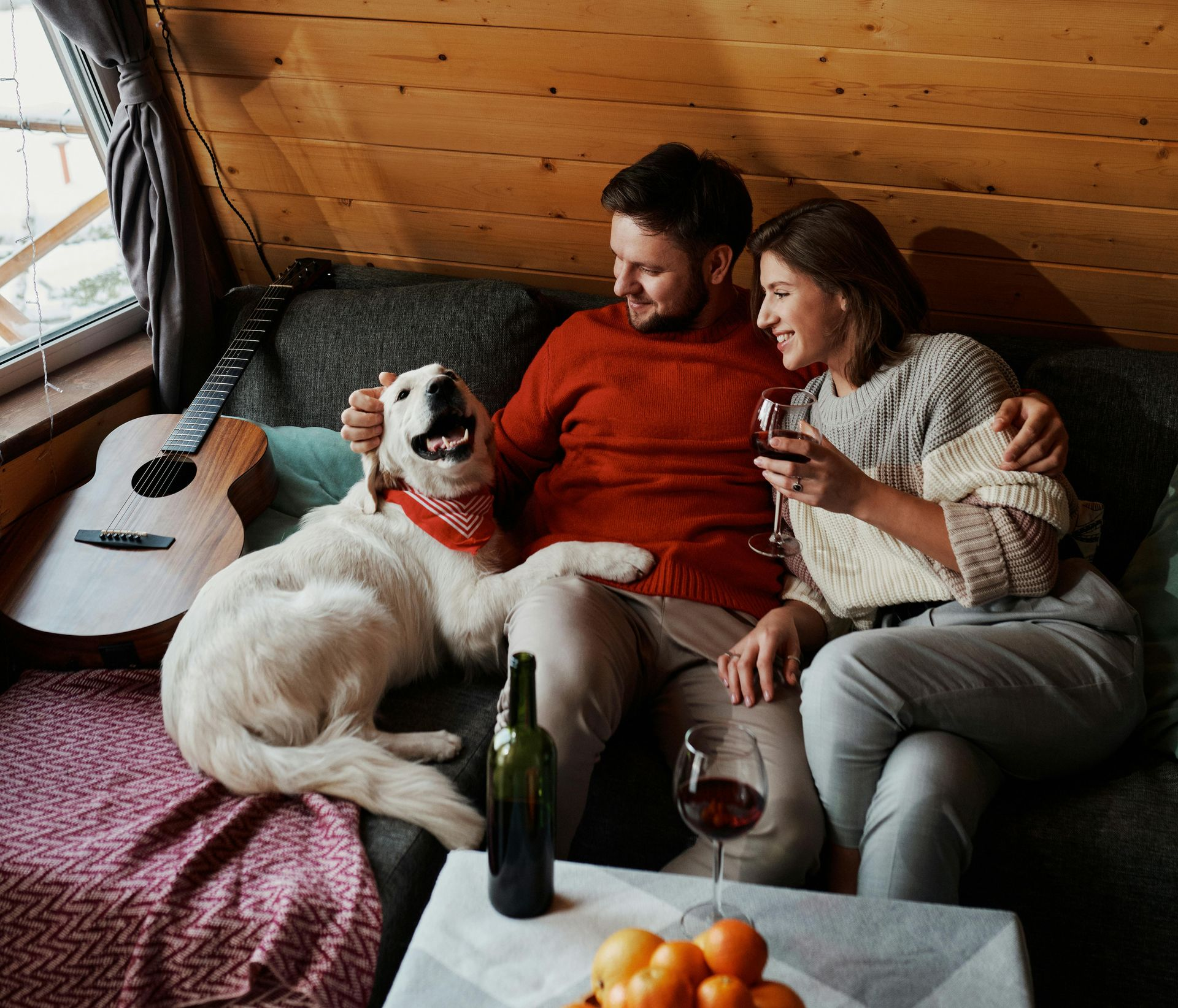 Couple on a couch with a dog and guitar, drinking wine in a cabin-like setting.