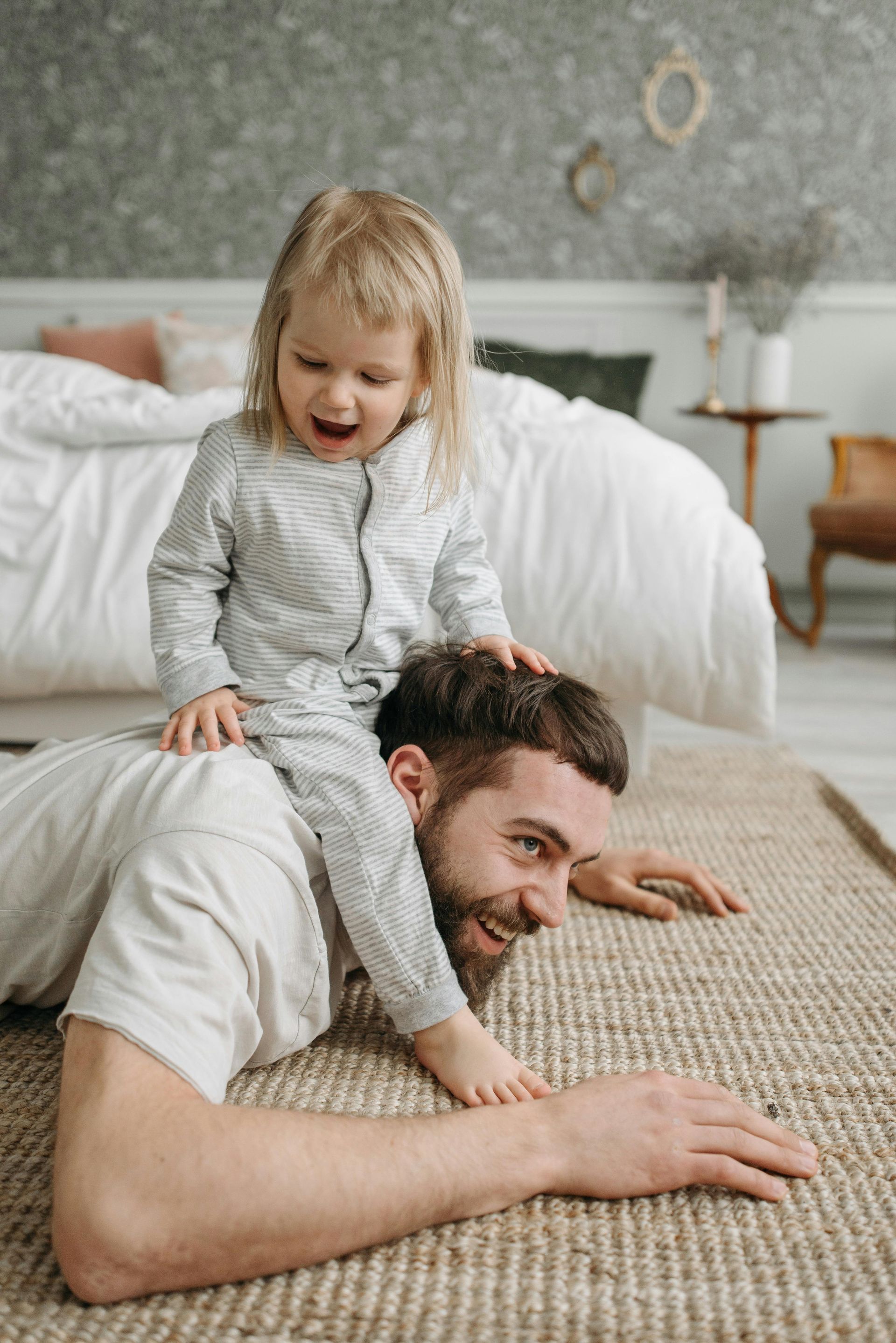 Man lying on a rug with a child sitting on his shoulders in a bedroom. The child is laughing.