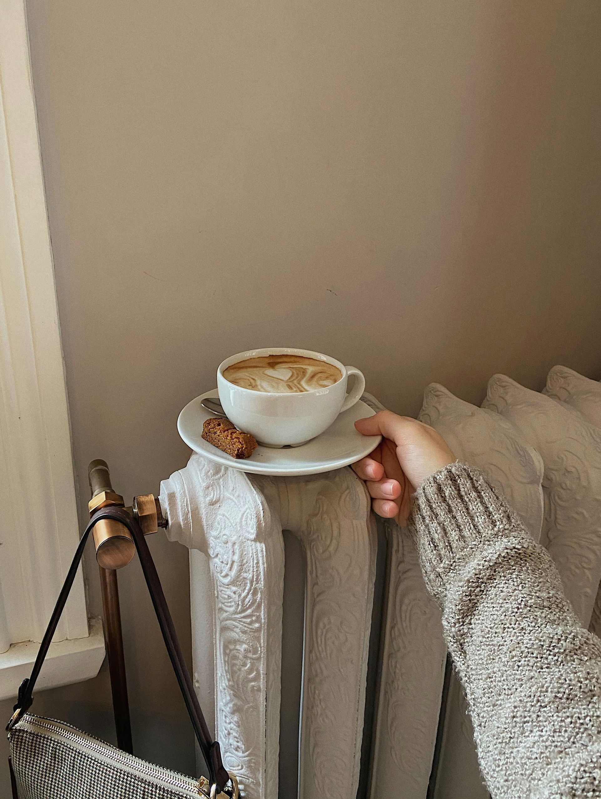 Hand holding a saucer with coffee and a cookie on a radiator, next to a purse.