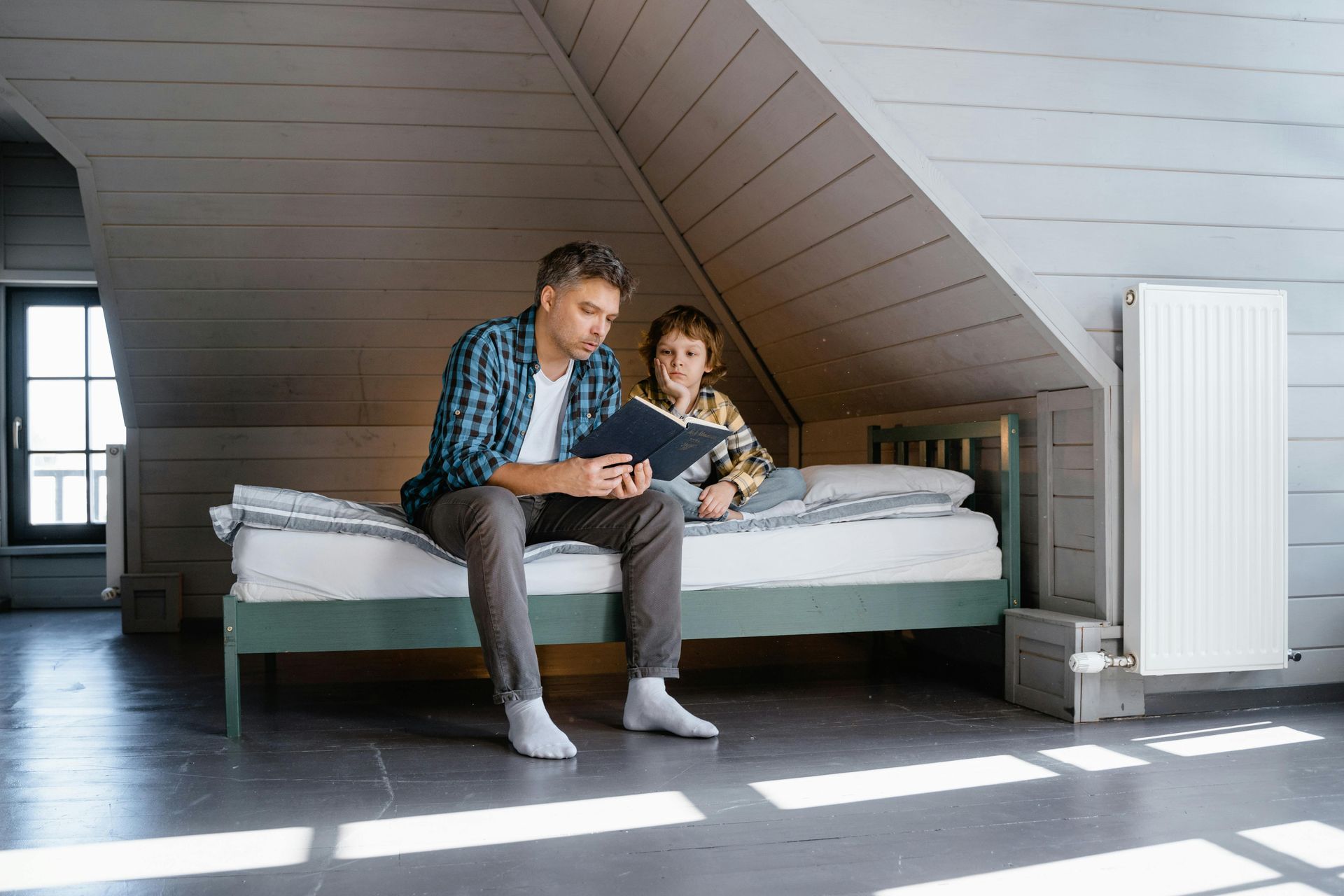 Man and child sitting on a bed in an attic, looking at a tablet.