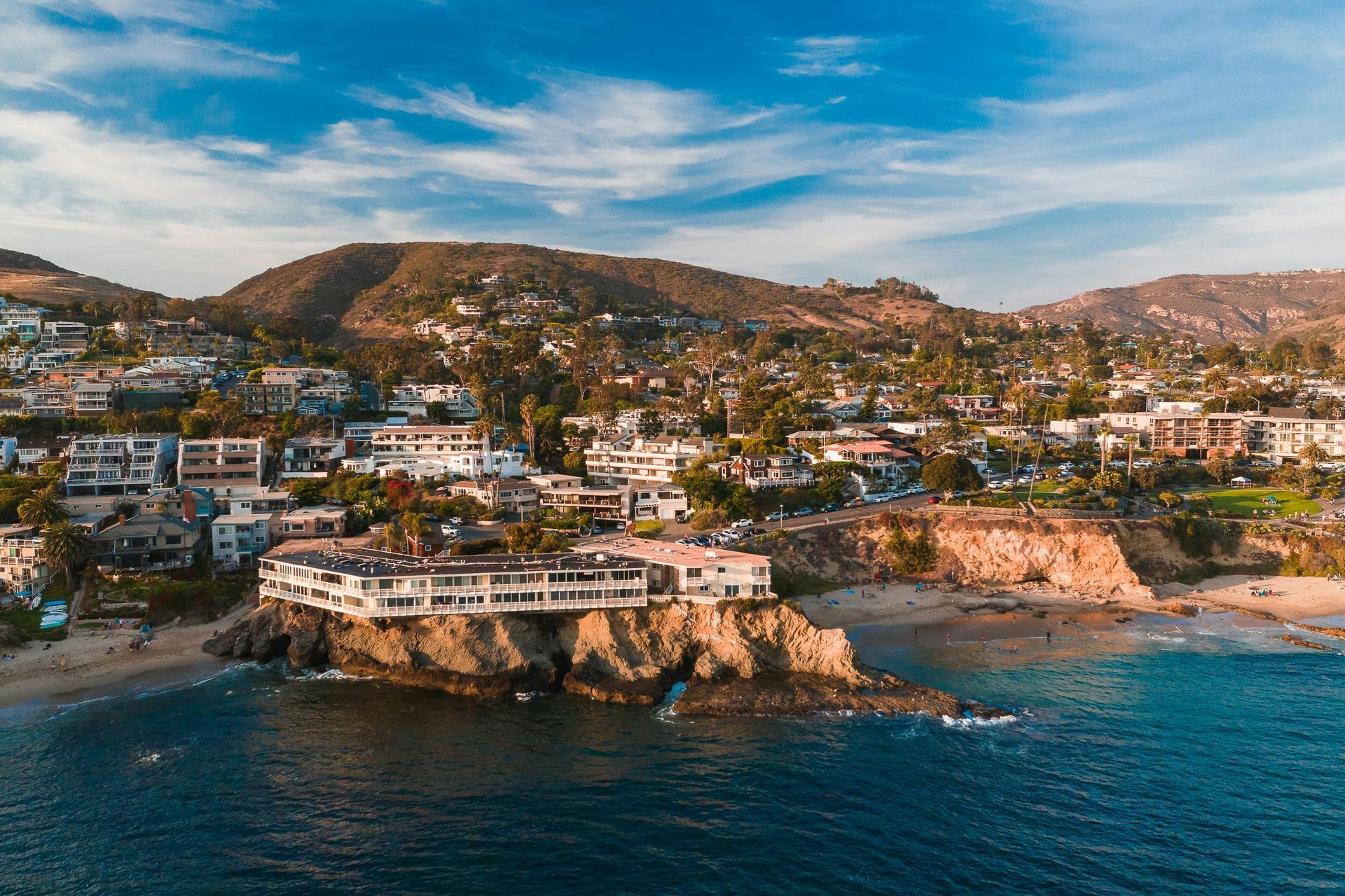 Coastal town with buildings on cliffs, a sandy beach, and a mountain backdrop; blue water and sky.