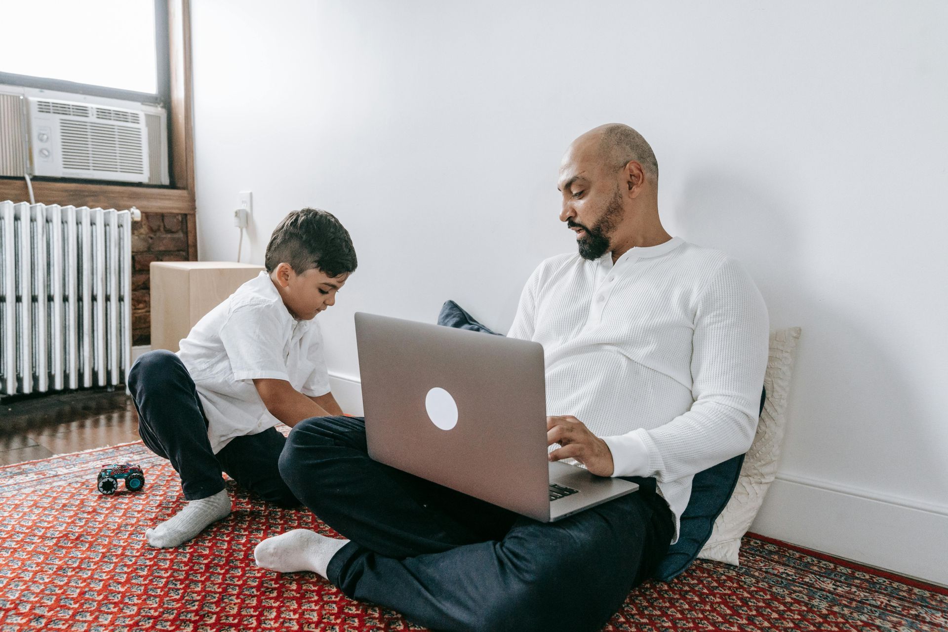 Man working on laptop, sitting on a rug with a child, in a room with a radiator.