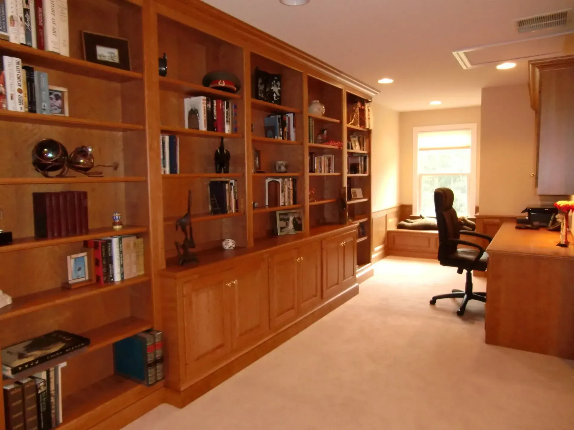 Built-in wooden bookshelves and cabinets in a home office. Light tan carpet and walls, desk, and office chair.