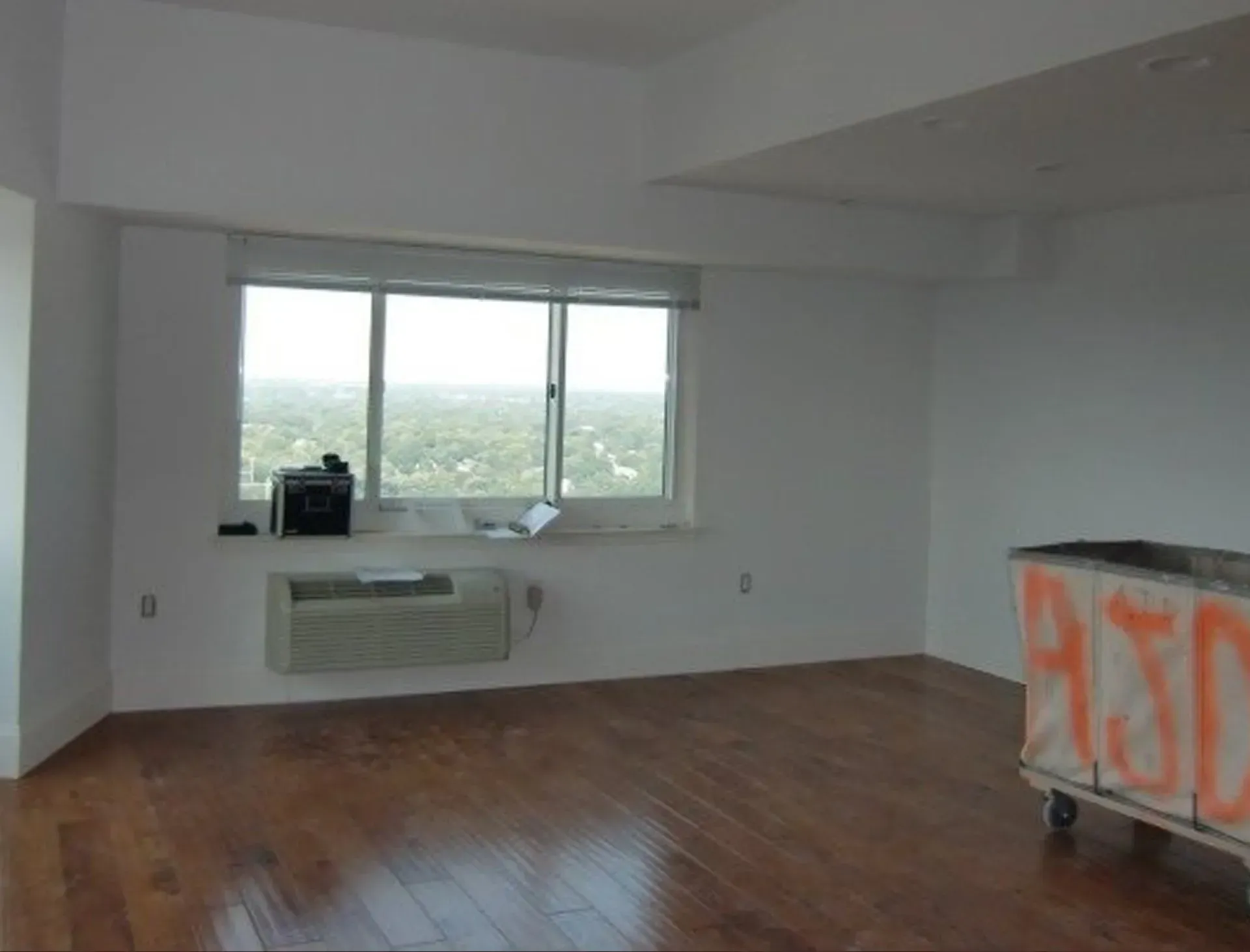 Empty room with hardwood floors, a window with a view, and a graffiti-covered cart.