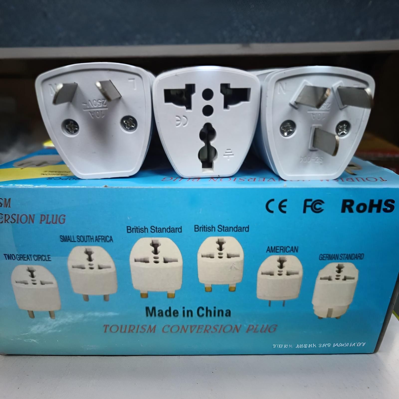 Tres enchufes adaptadores de viaje blancos en una caja azul, que muestran diferentes tipos de enchufes; "Hecho en China".