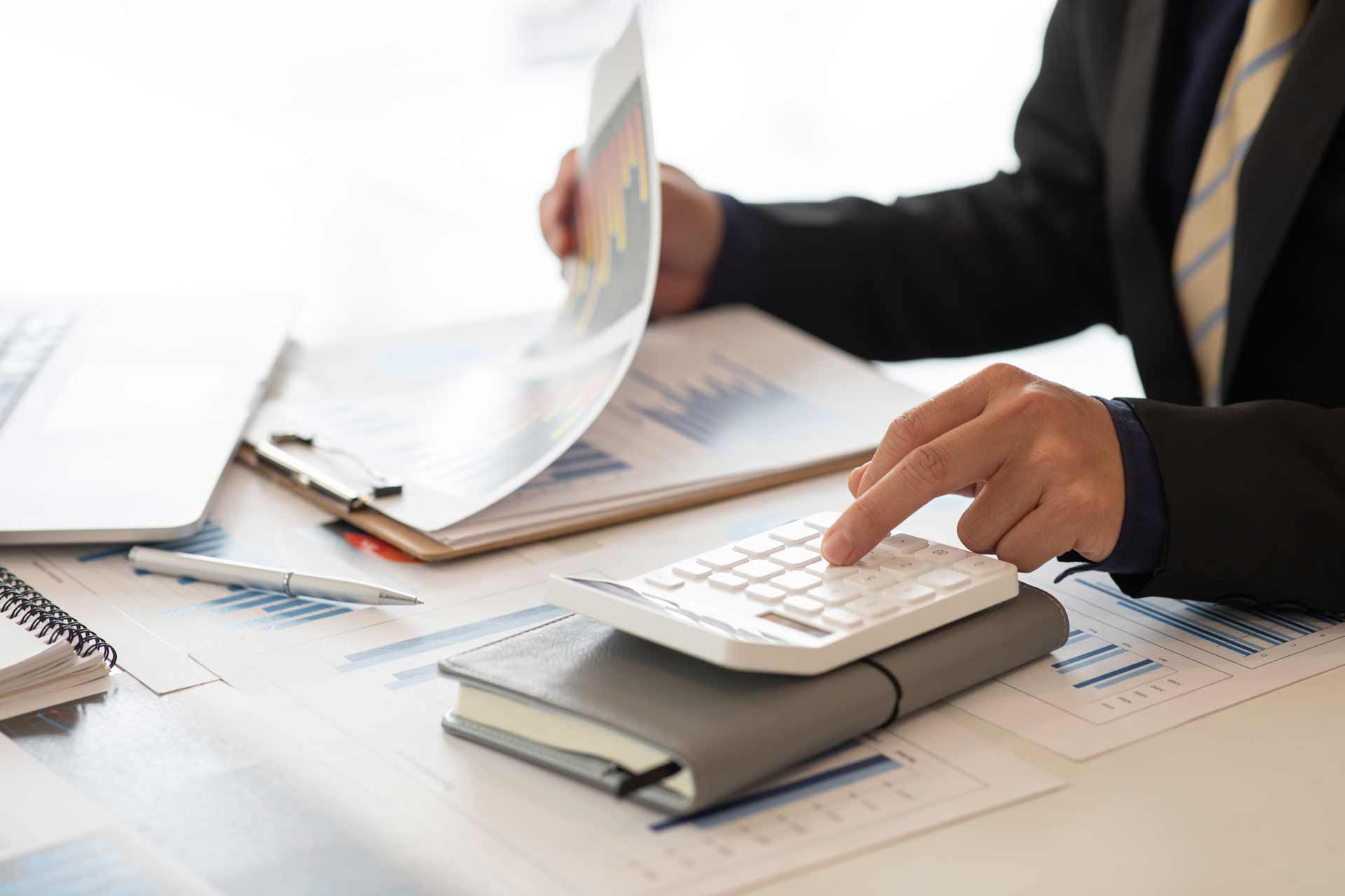 Person in suit calculating figures with a calculator, reviewing charts and data on a desk