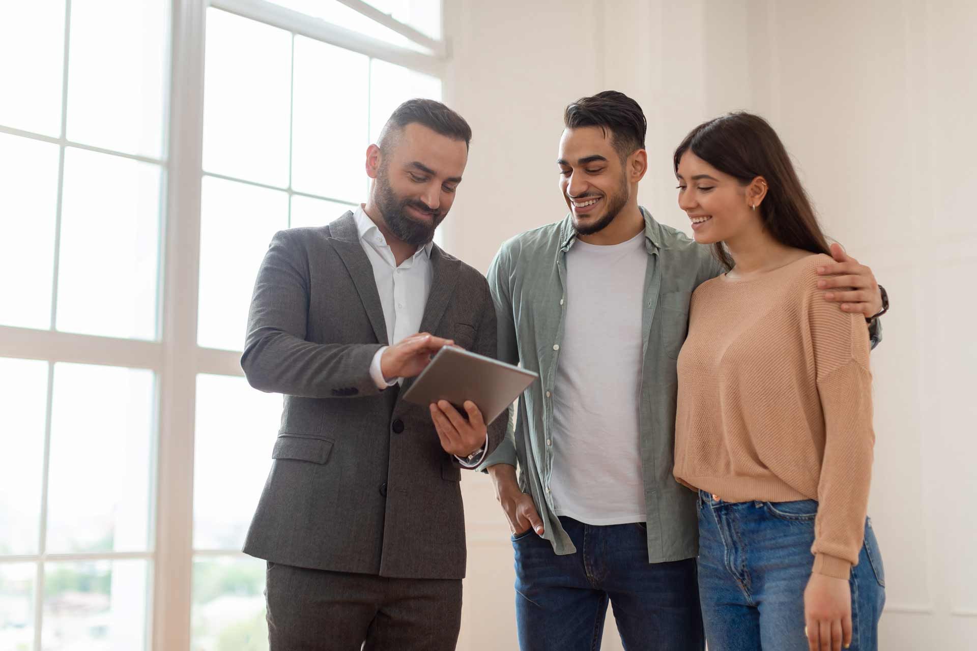 Real estate agent showing couple a tablet in a bright, empty room. They are smiling, windows in background.