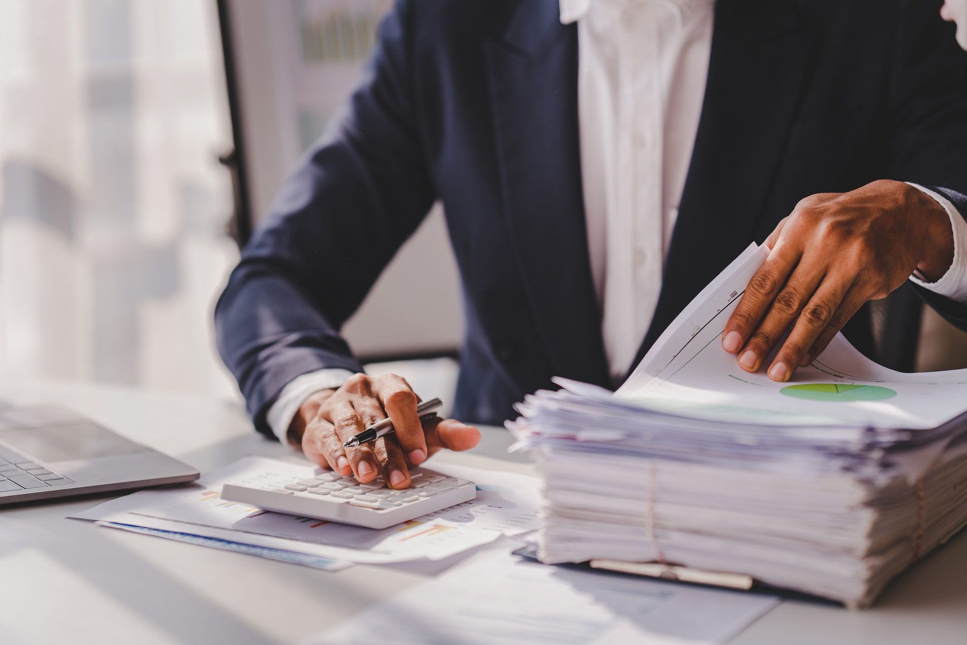 Person in suit using a calculator at a desk, reviewing documents and paperwork.