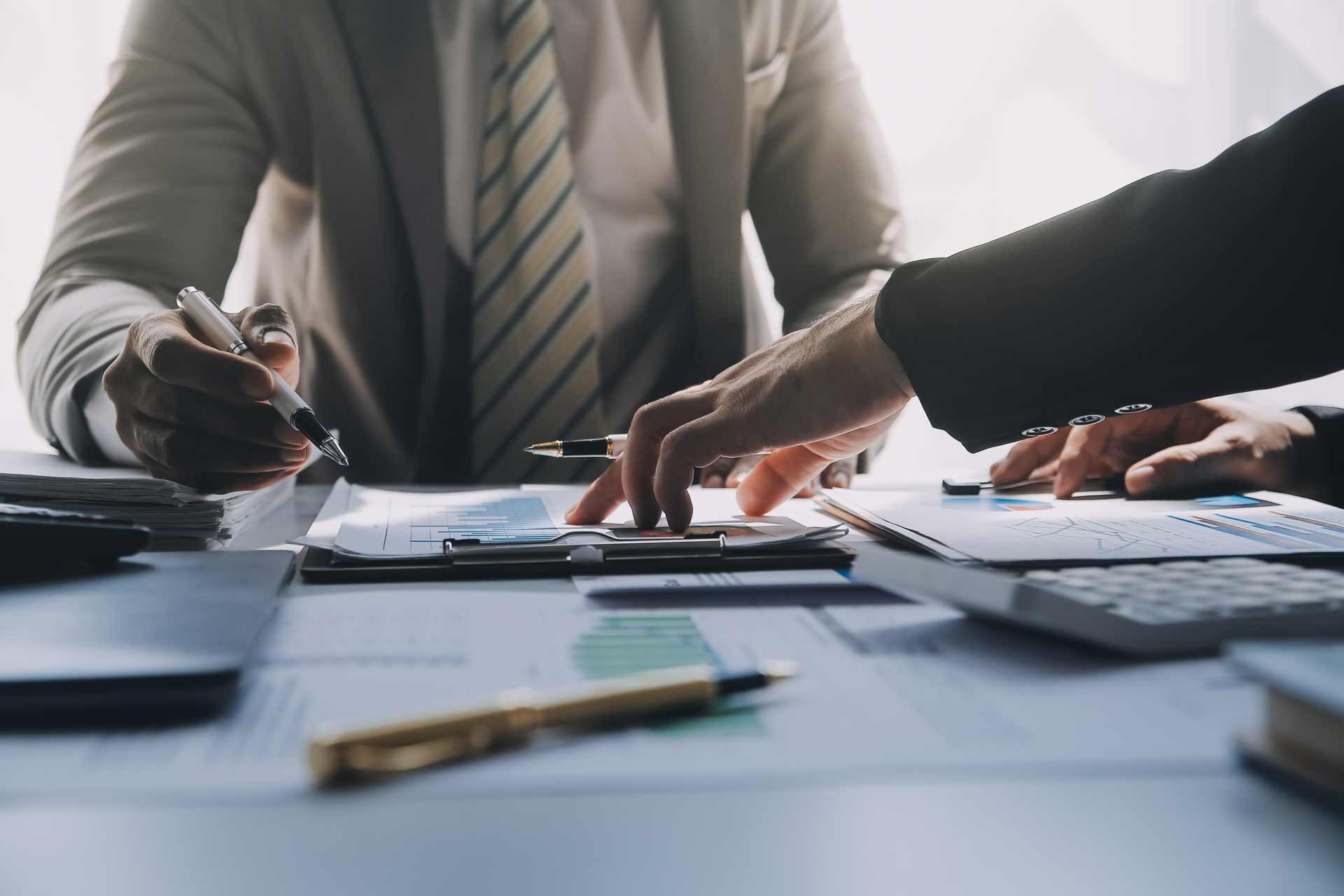 Two businesspeople review documents, hands pointing, wearing suits, at a desk with calculator and pen
