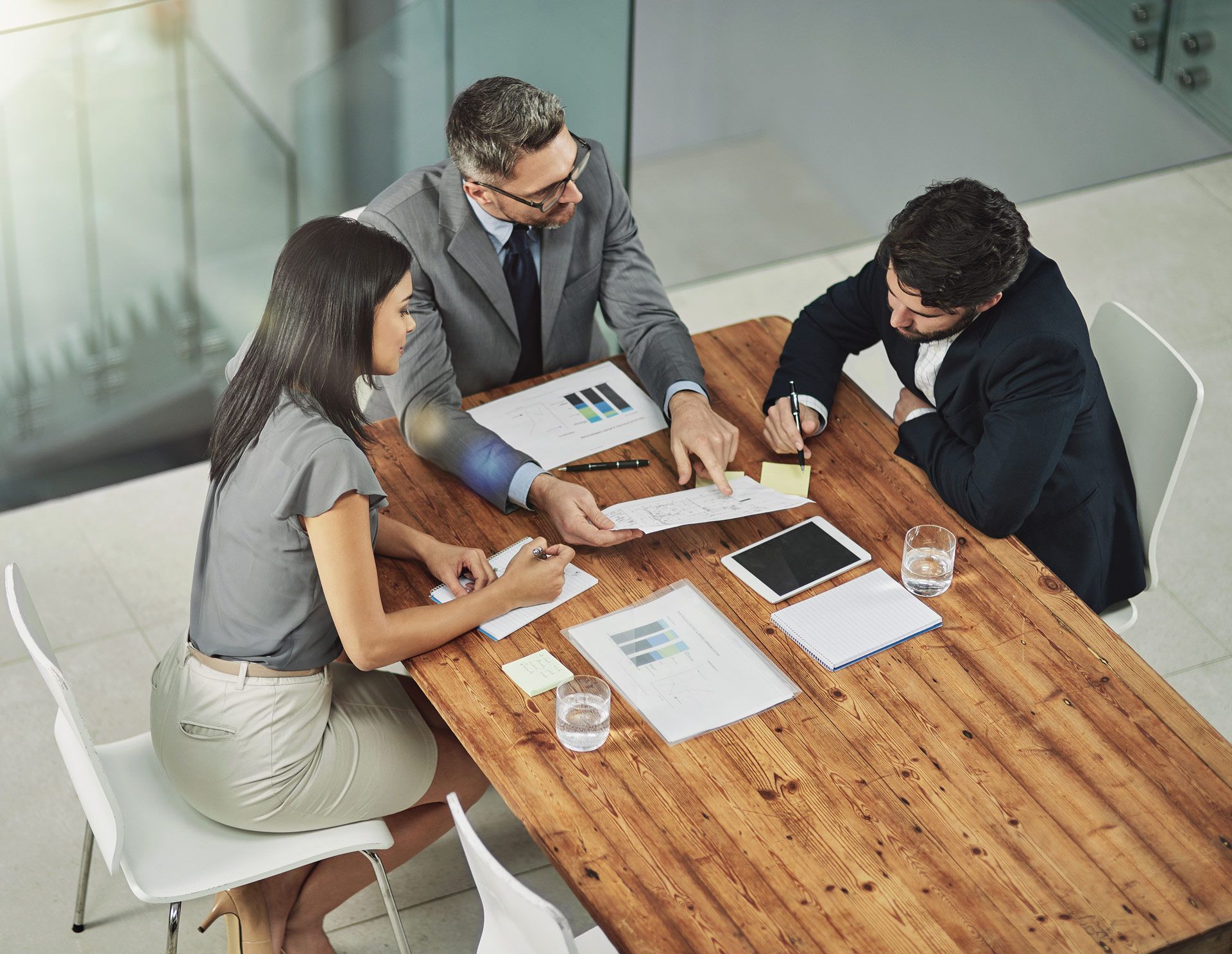 Three people in business attire reviewing documents at a wooden table, in a modern office.