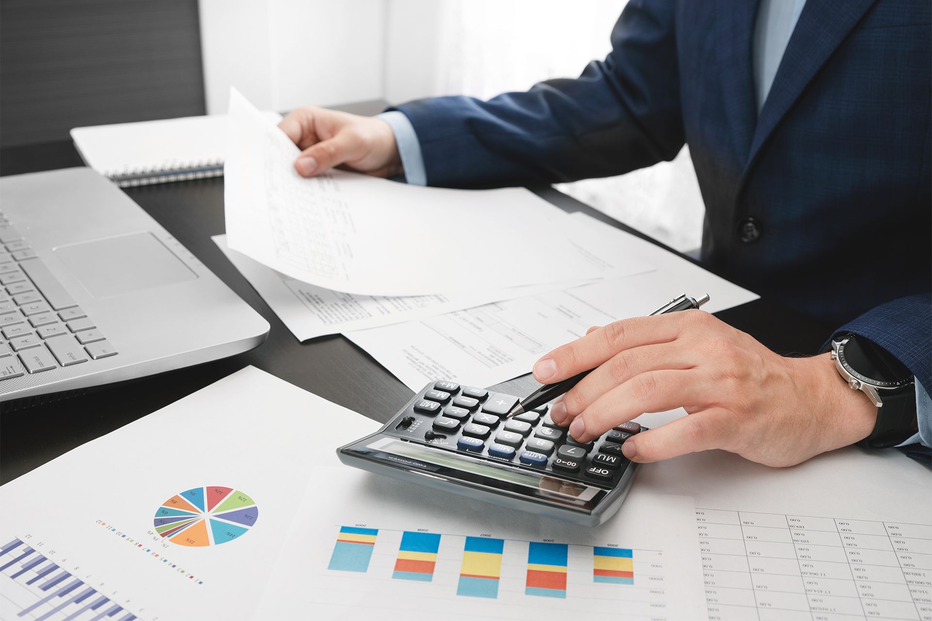 Person in suit using a calculator, reviewing papers and financial charts at a desk.