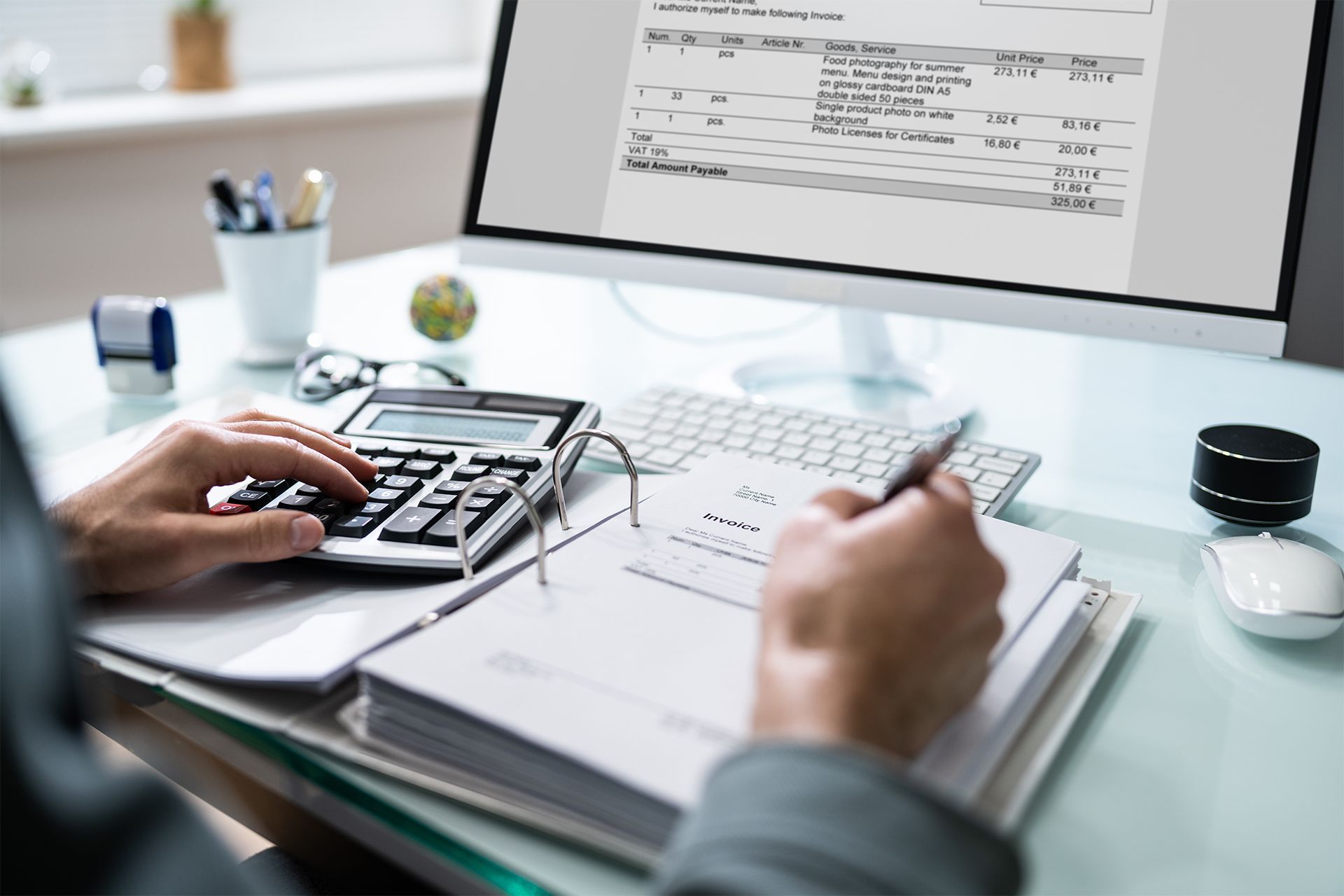 Person using a calculator and pen while reviewing a document and computer screen at a desk.