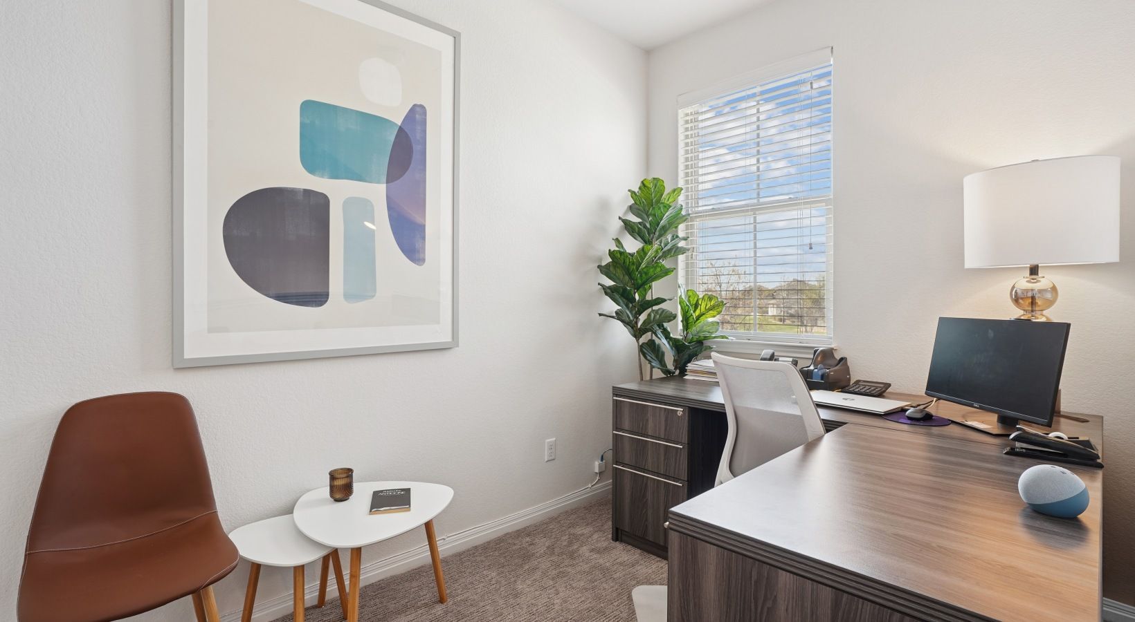 Interior apartment office with desk, chair, monitor, lamp, plant, and abstract wall art near a window.
