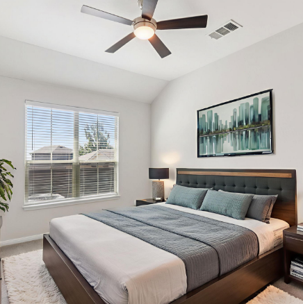 Bedroom with bed, art, window, and ceiling fan. White walls, grey bedding, and neutral tones.