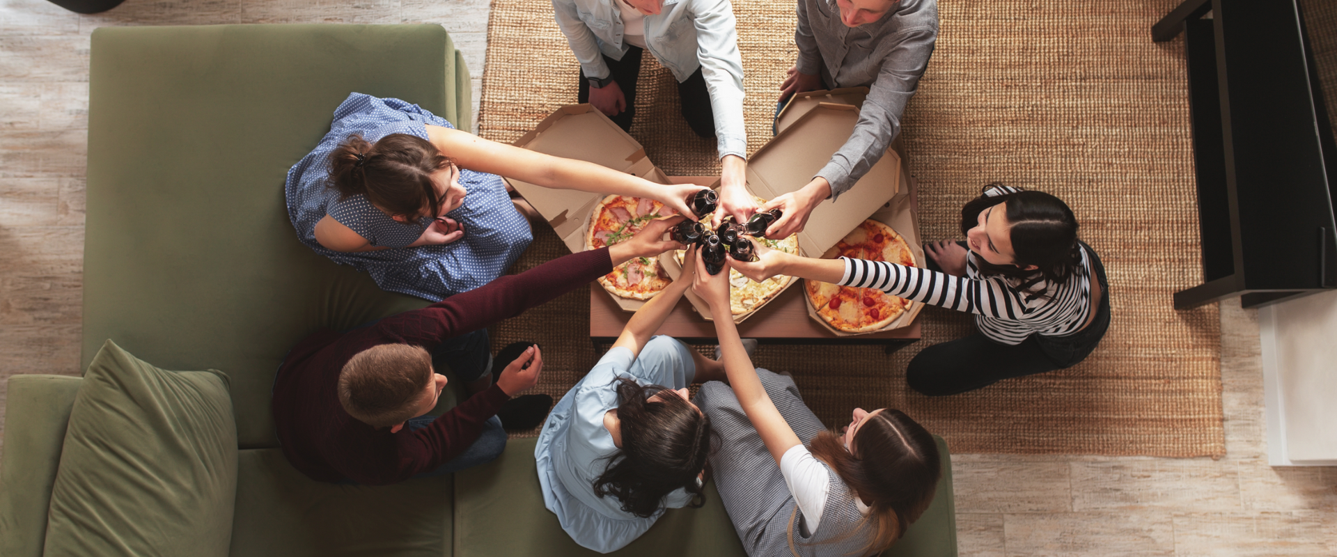 A group of people sitting around pizzas and toasting with dark beverages, viewed from above.