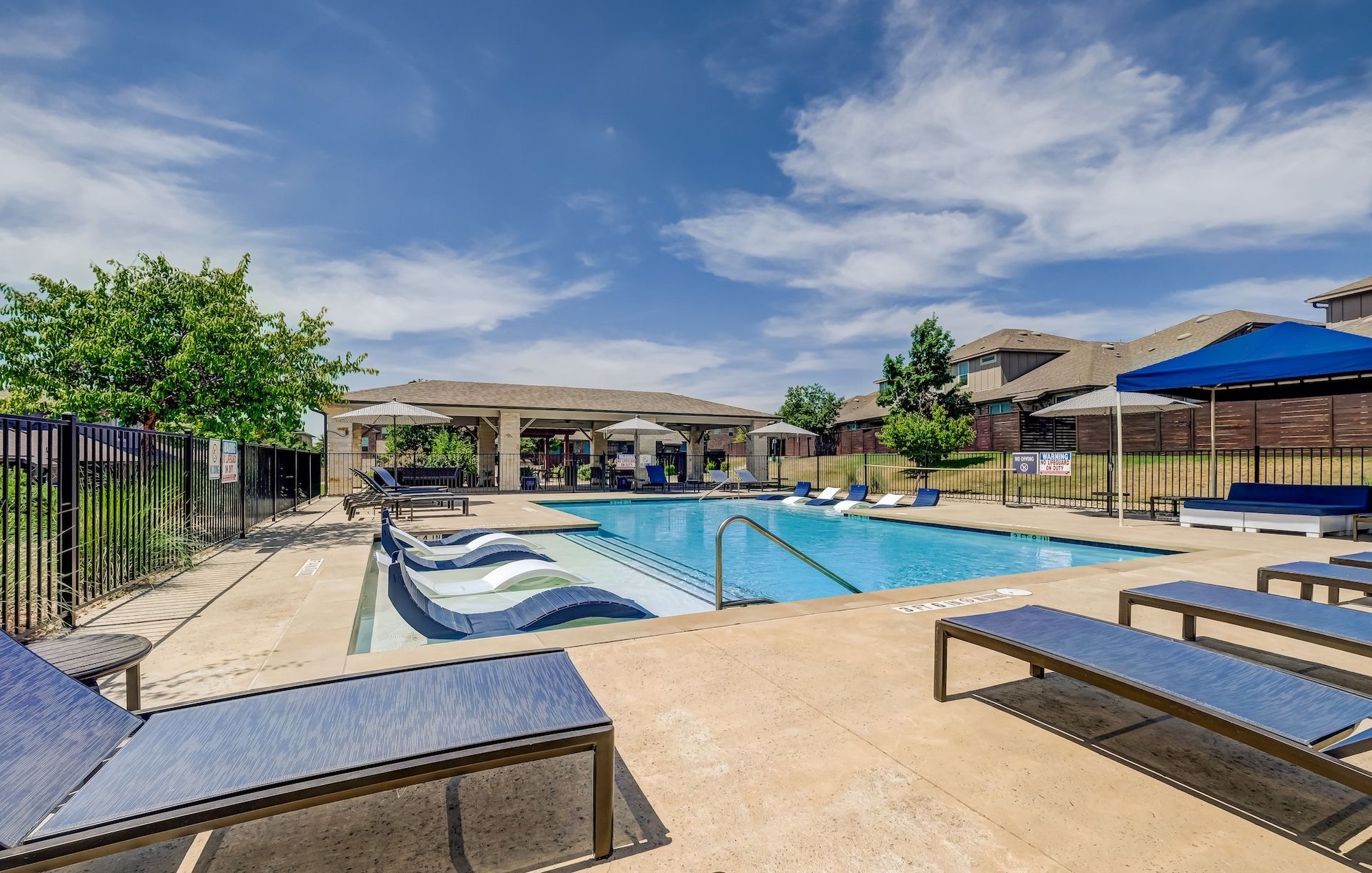 Pool area with blue lounge chairs, pool, and a shaded structure under a blue sky.