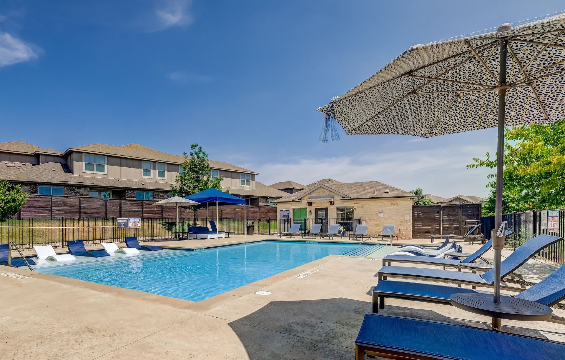 Pool area with lounge chairs, umbrella, and buildings under a blue sky.