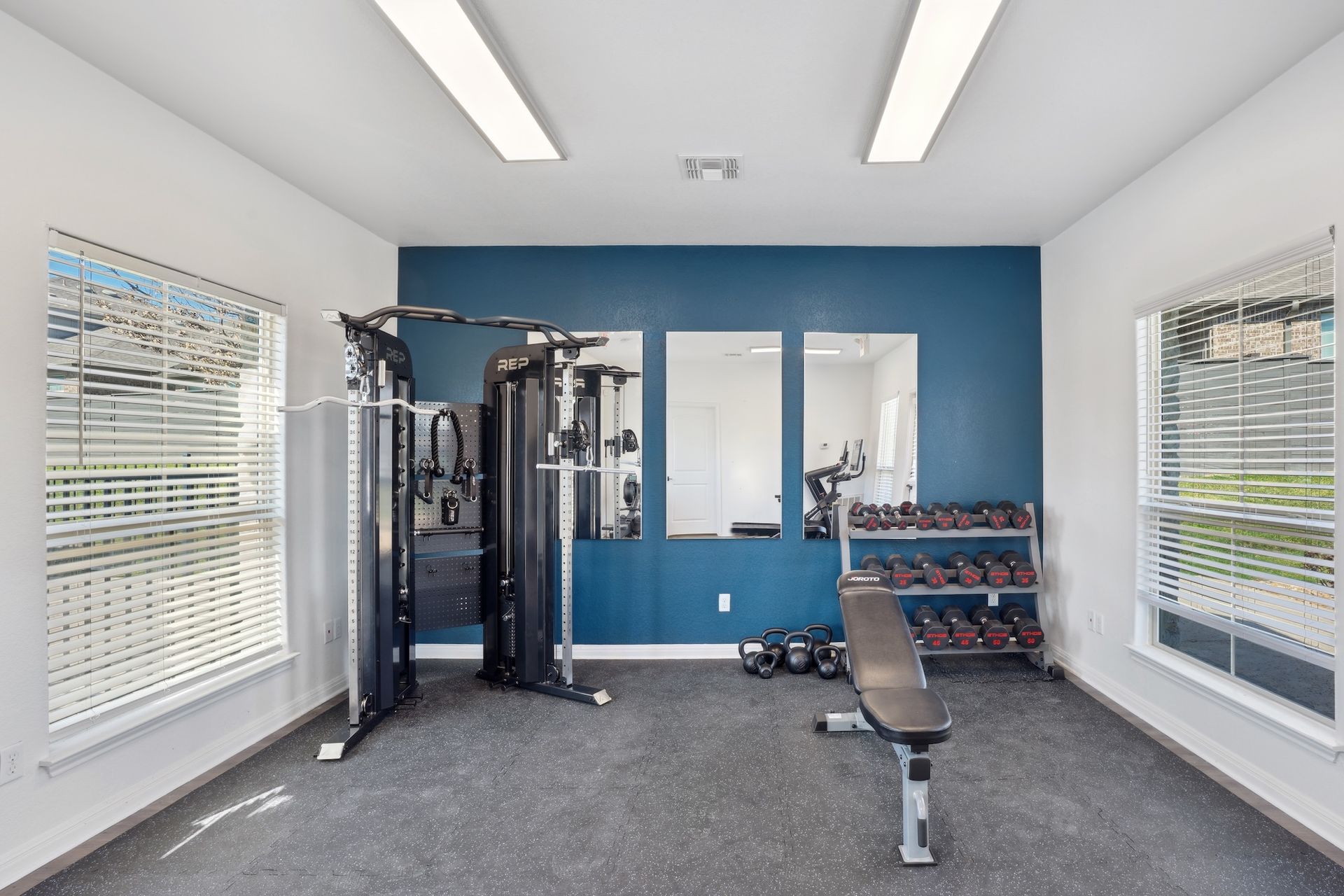 Gym with blue accent wall, weight machines, dumbbells, weight bench, mirrors, and windows.