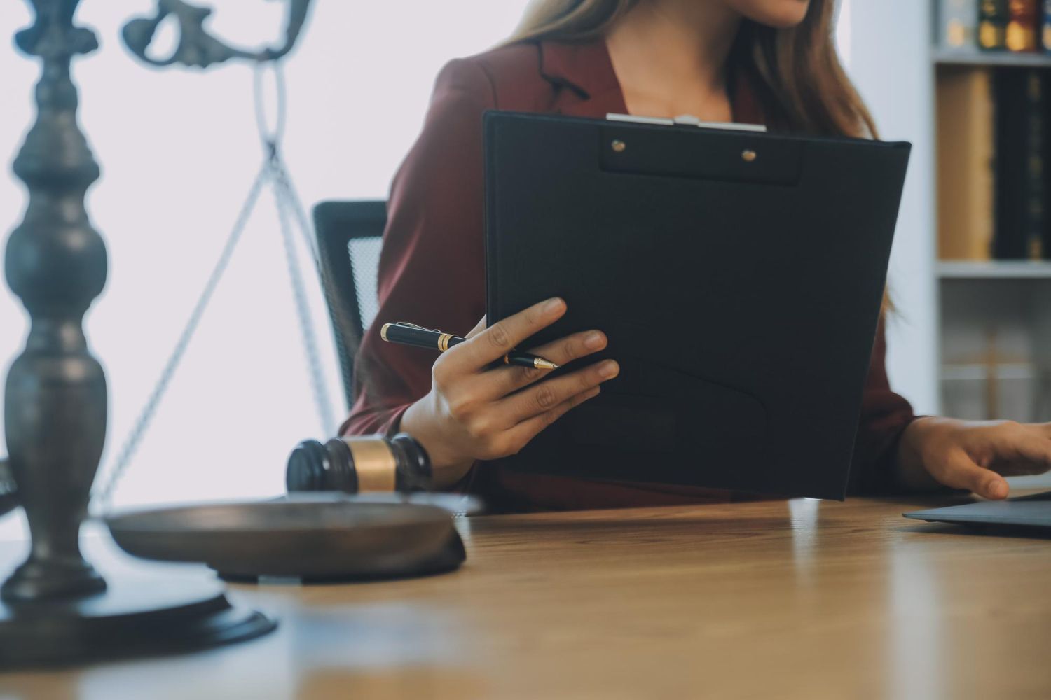 Woman in a burgundy blazer holding a clipboard, a gavel, and a laptop in a law office setting.