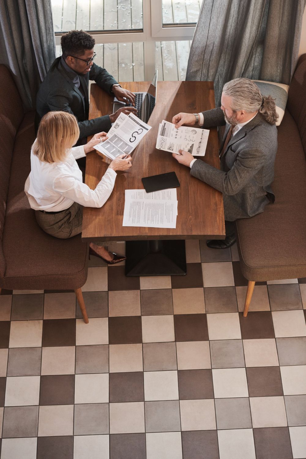 Business team reviewing documents at a table. Wooden table with people, papers, and laptop. Checkered floor.